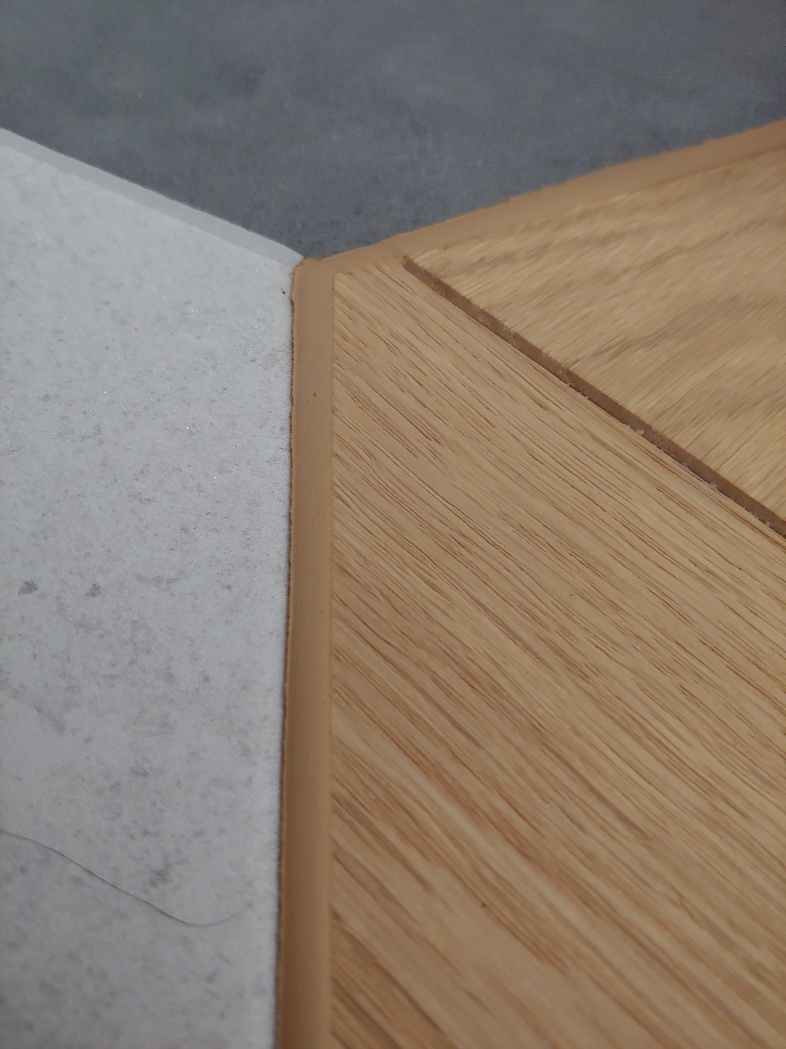 Zbliżenie na styk jasnego parkietu ułożonego w jodełkę z szarą, porowatą płytką, połączonych beżową listwą przypodłogową.