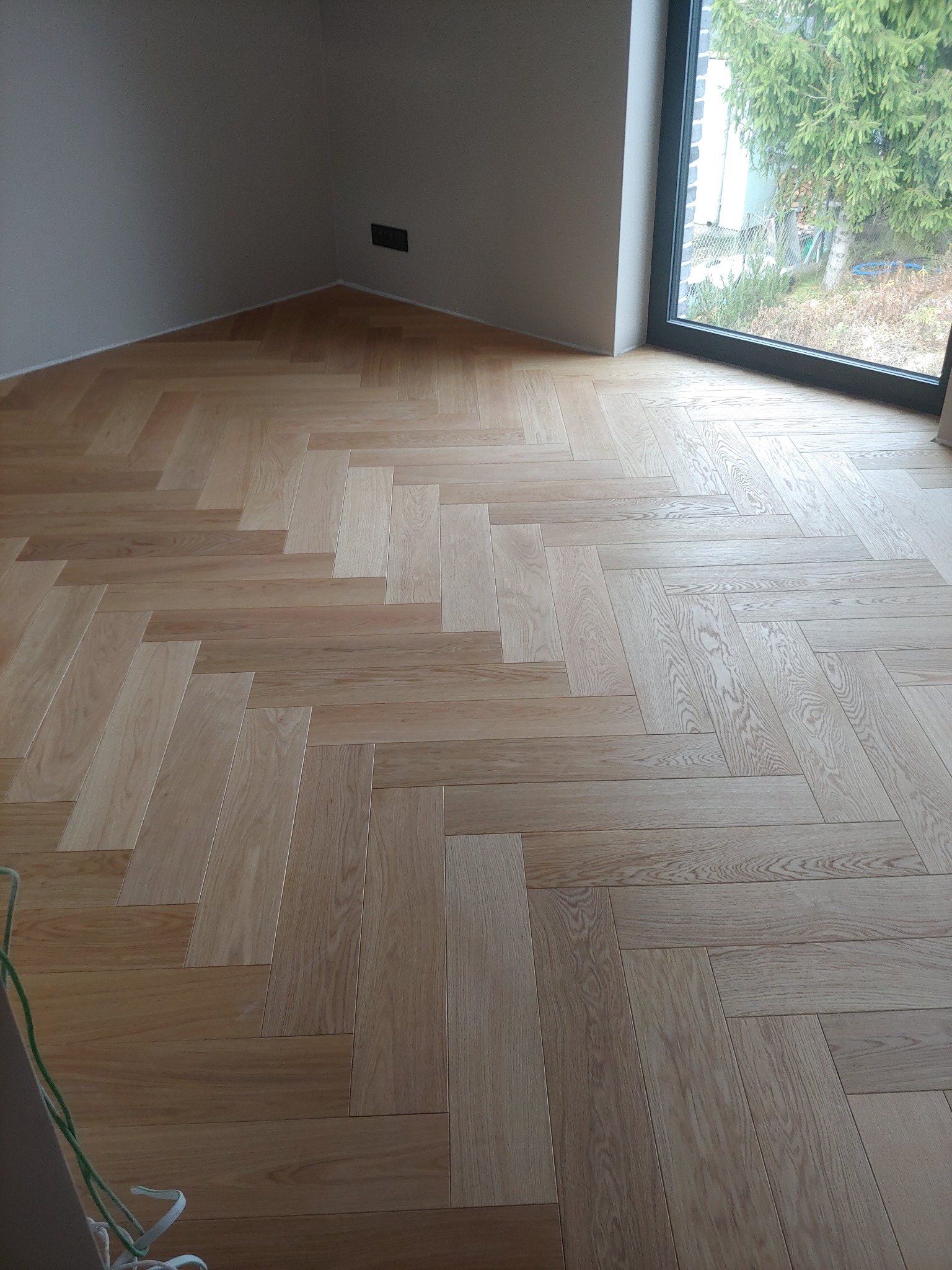 Jasny parkiet ułożony we wzór jodełki francuskiej w rogu pokoju z oknem wychodzącym na zieleń.