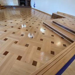 Mparkiet - Świeżo położony, błyszczący parkiet w geometryczny wzór z jasnego drewna z ciemnobrązowymi akcentami, widoczne listwy przypodłogowe w niebieskiej ochronnej taśmie malarskiej.