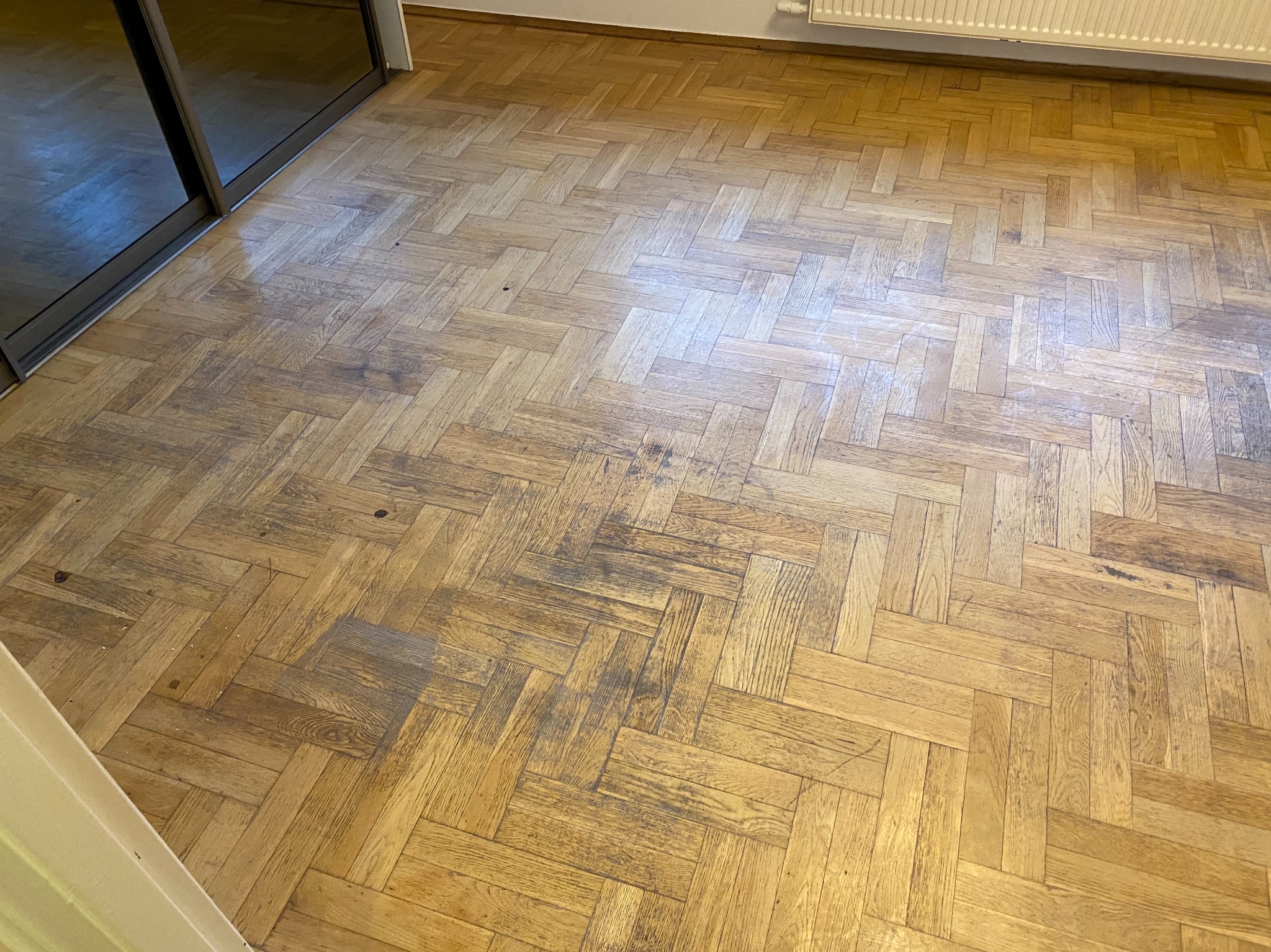 Parkiet ułożony w jodełkę, z widocznymi śladami użytkowania i zarysowaniami, przed renowacją, w tle grzejnik i przesuwne drzwi.