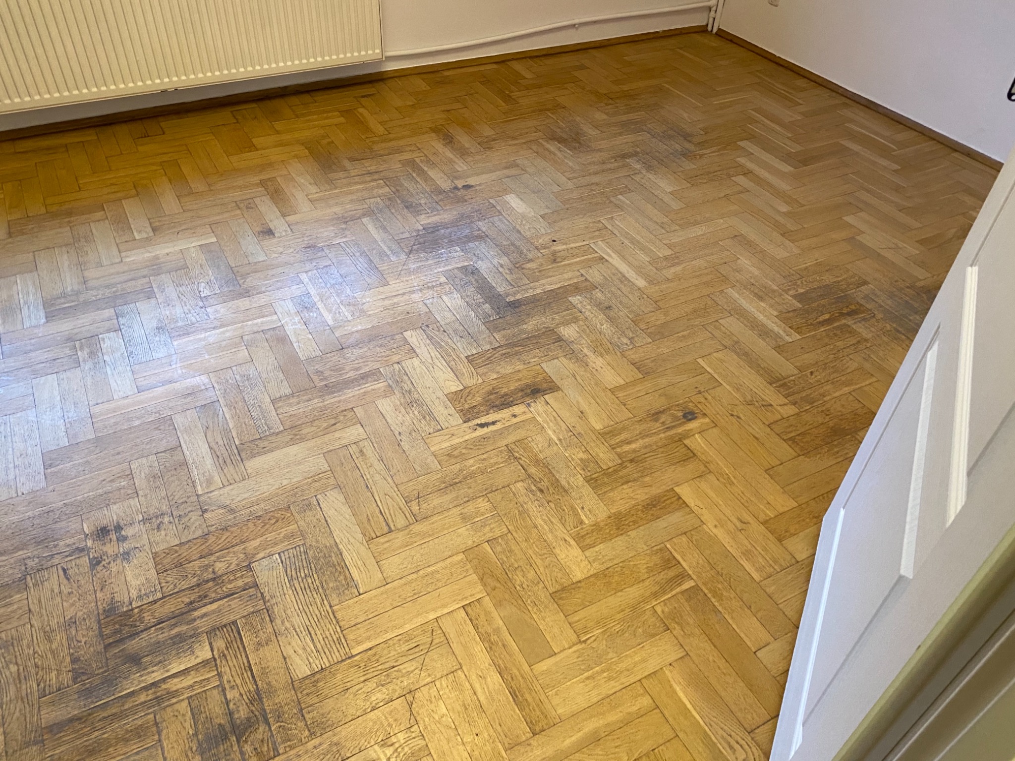 Parkiet ułożony w jodełkę, widoczne ślady użytkowania i przebarwienia, oświetlenie dzienne, fragment grzejnika i drzwi w tle.