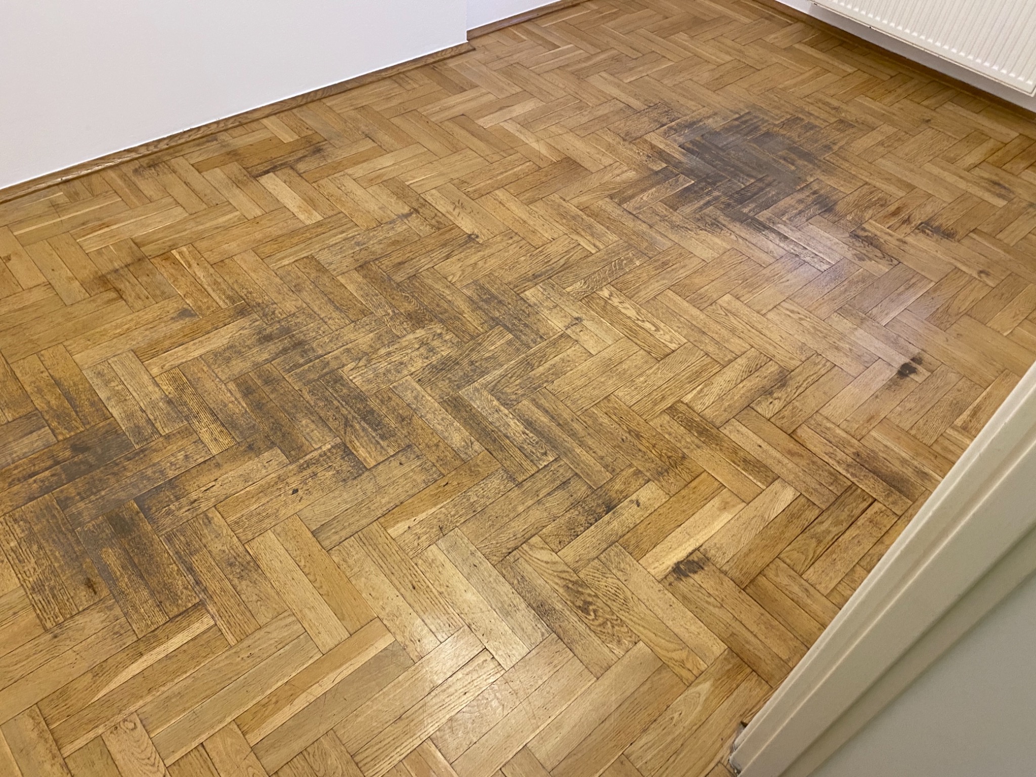 Parkiet ułożony w jodełkę, z widocznymi ciemnymi plamami i śladami użytkowania, wymagający renowacji. Widoczne fragmenty białej ściany i listwy przypodłogowej.