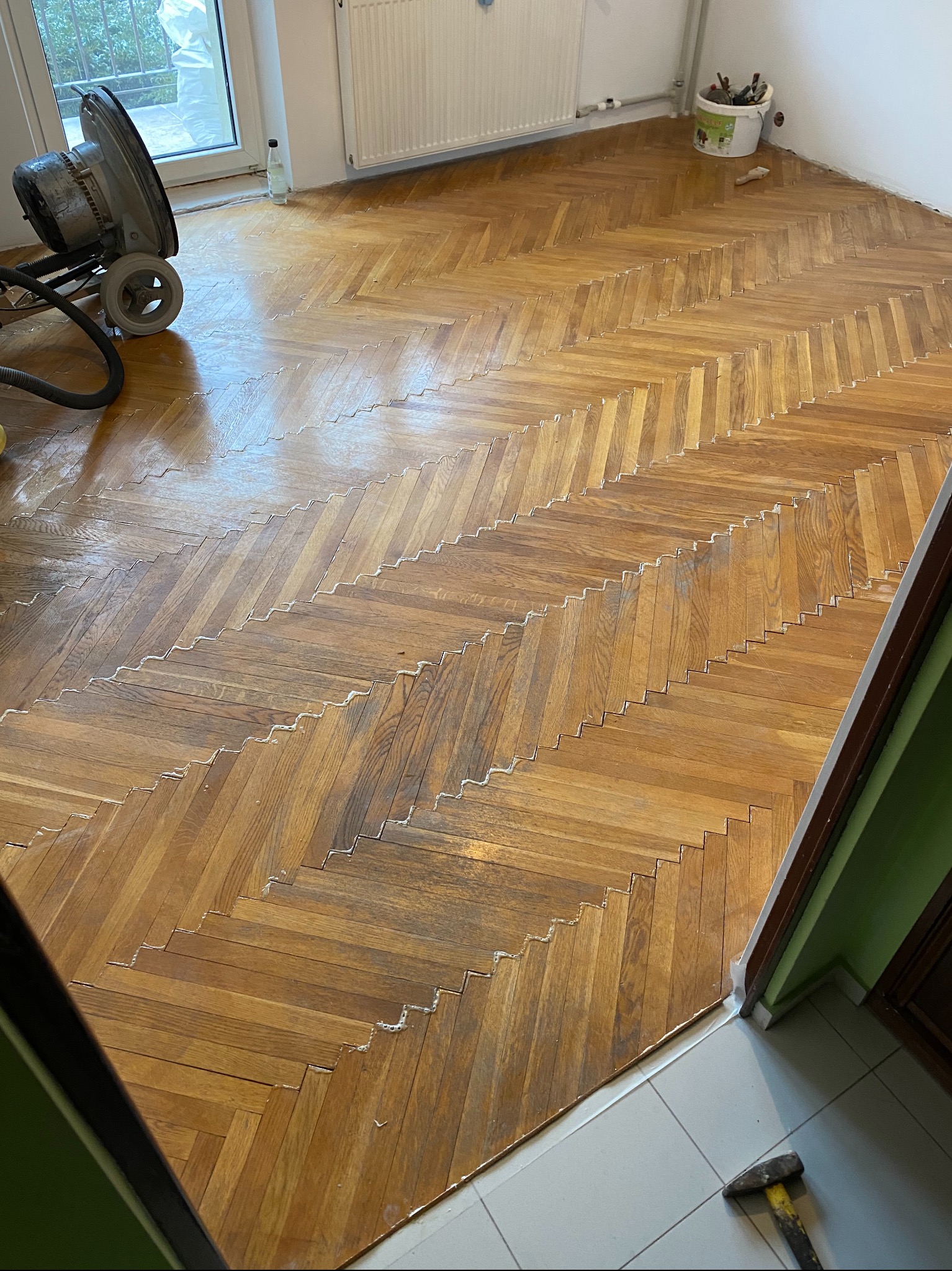 Parkiet ułożony w jodełkę, częściowo pokryty warstwą lakieru, obok maszyna do szlifowania i wiadro z narzędziami malarskimi.