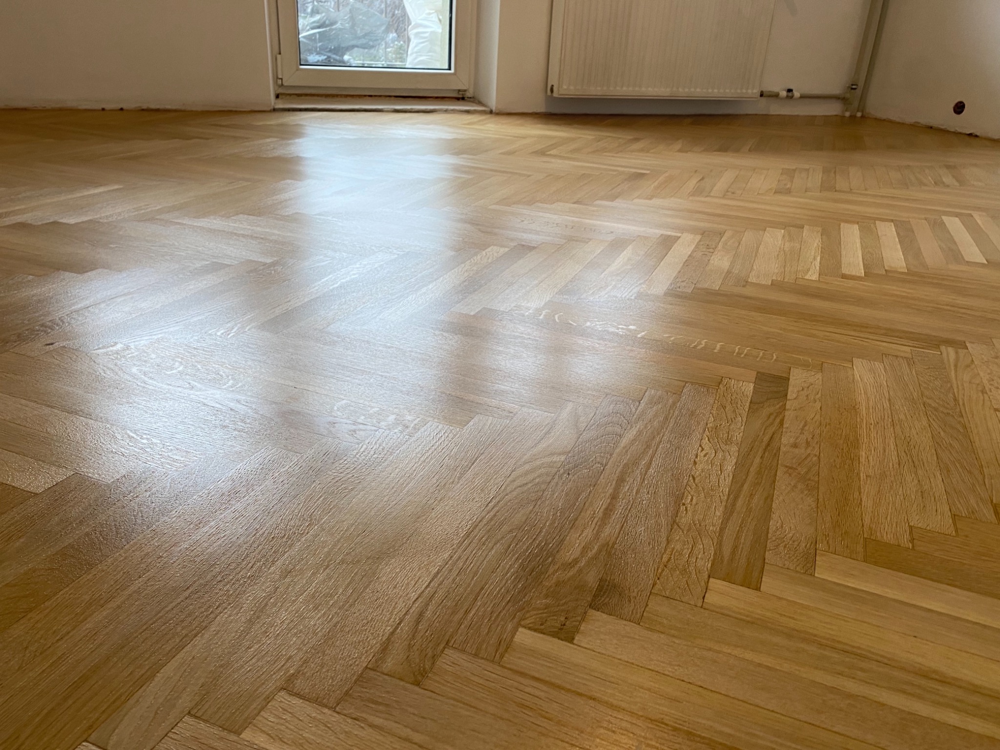 Ułożony parkiet w jodełkę w jasnym, pustym pokoju z grzejnikiem i oknem w tle.