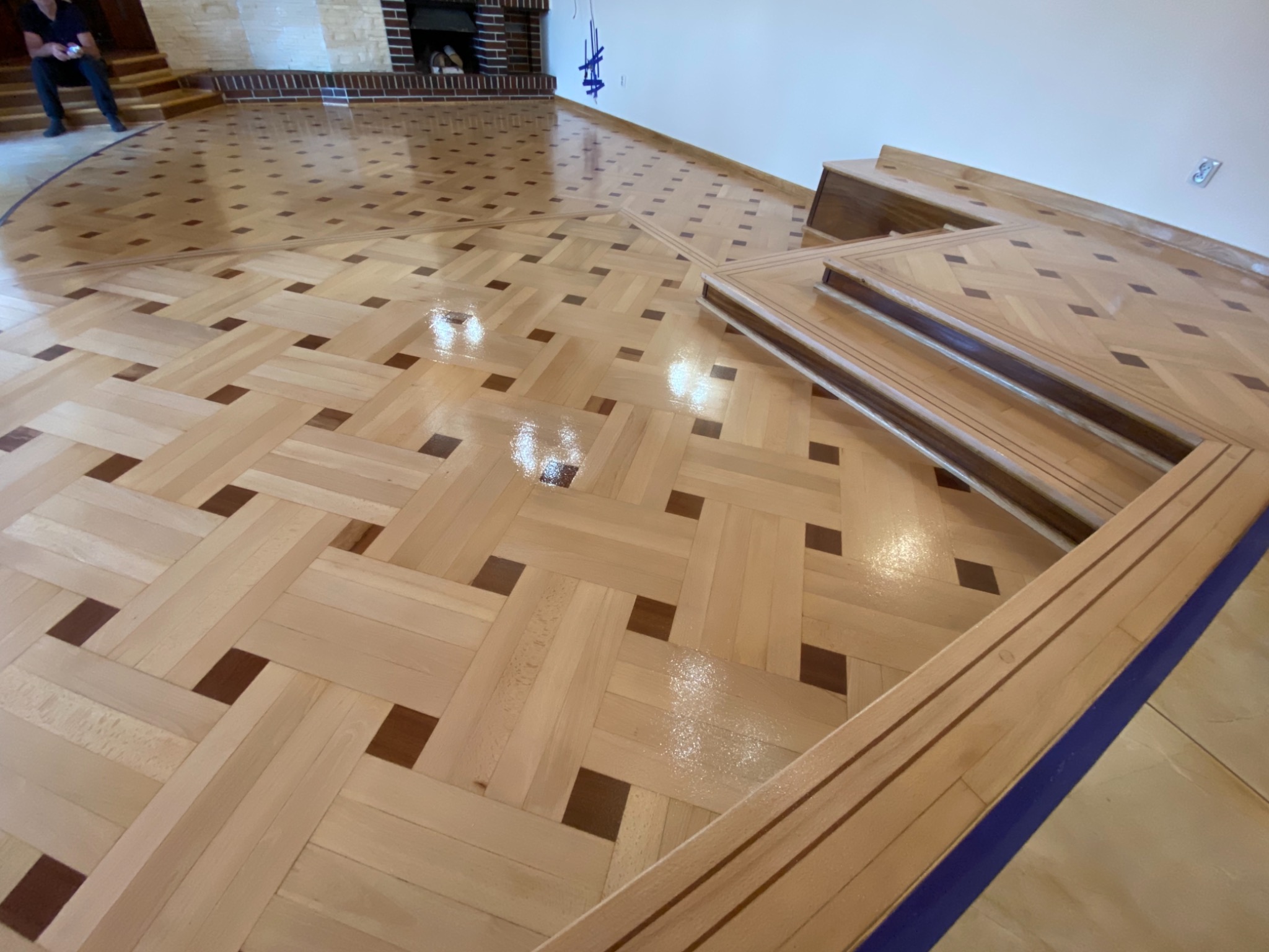 Świeżo położony, błyszczący parkiet w geometryczny wzór z jasnego drewna z ciemnobrązowymi akcentami, widoczne listwy przypodłogowe w niebieskiej ochronnej taśmie malarskiej.