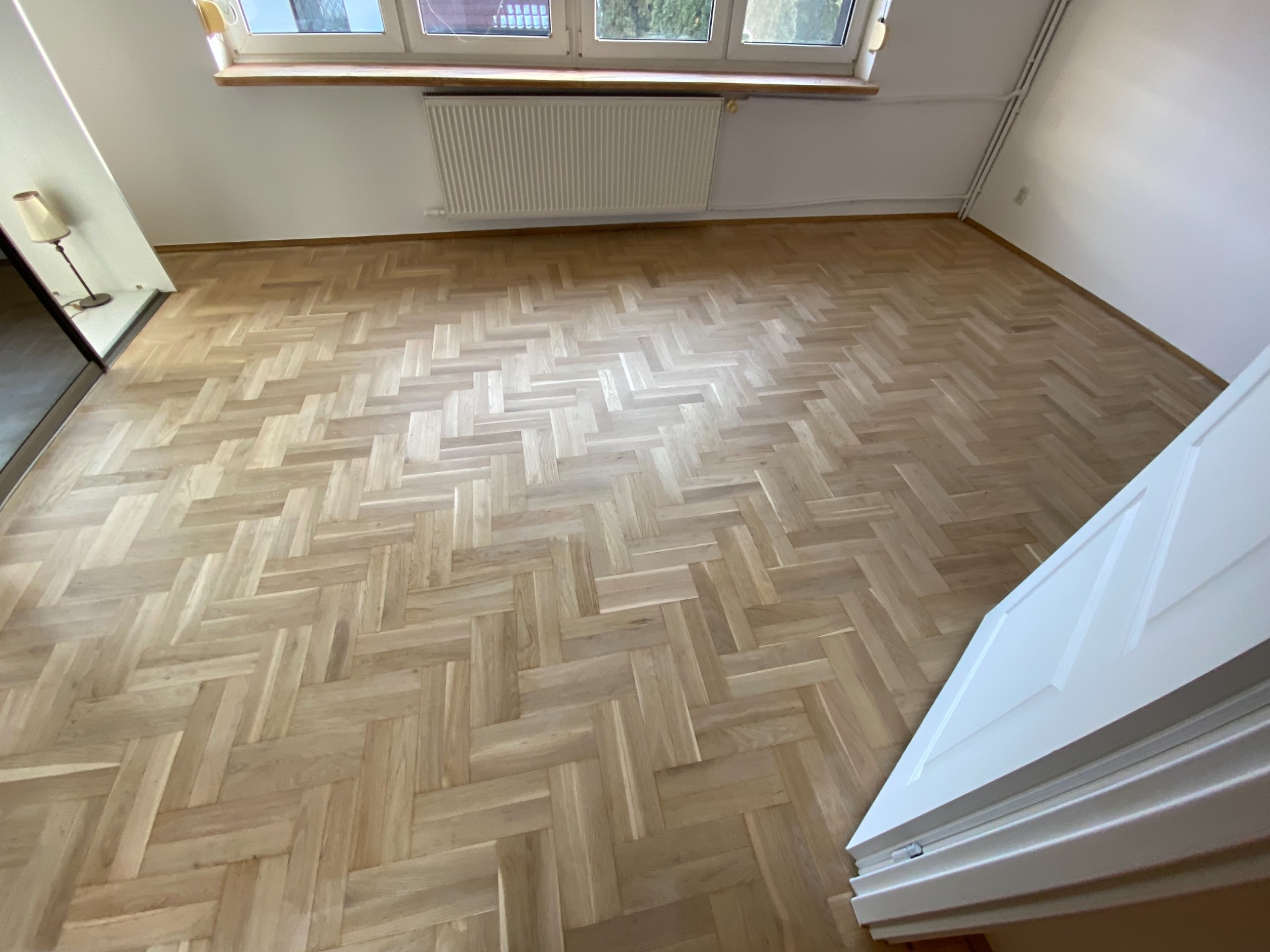 Jasny parkiet ułożony we wzór jodełki francuskiej w pustym pokoju z białymi ścianami i grzejnikiem pod oknem.