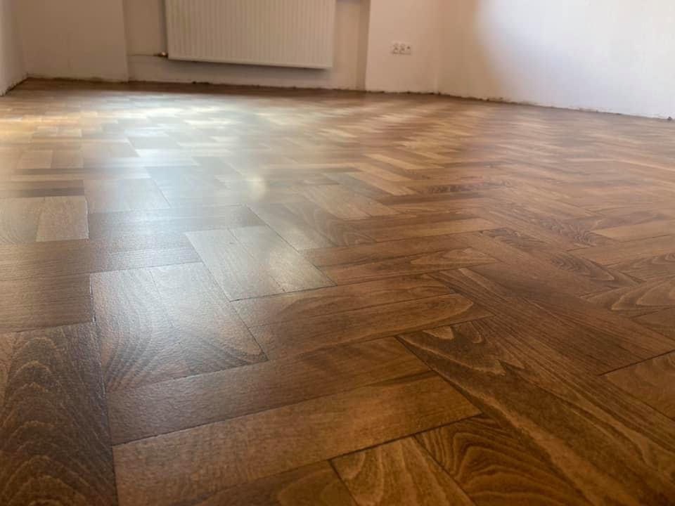 Odnowiona drewniana podłoga ułożona w jodełkę, z widocznym połyskiem lakieru i fragmentem grzejnika na ścianie w tle.