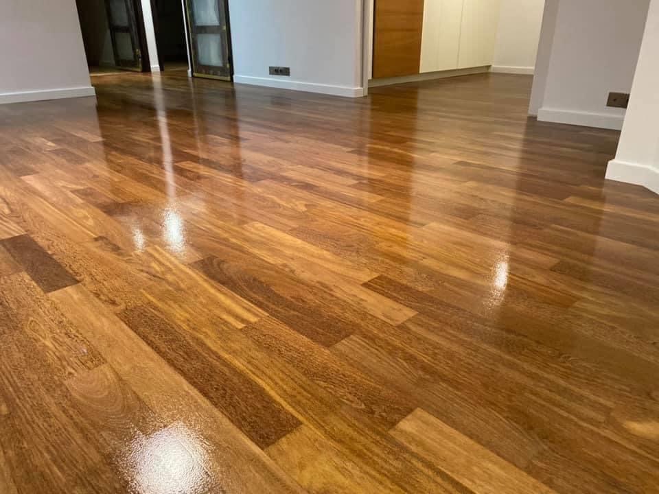 Błyszcząca, świeżo wycyklinowana podłoga parkietowa z wyraźnym usłojeniem, odbijająca światło w przestronnym, jasnym wnętrzu z białymi ścianami i ciemnymi drzwiami w tle.