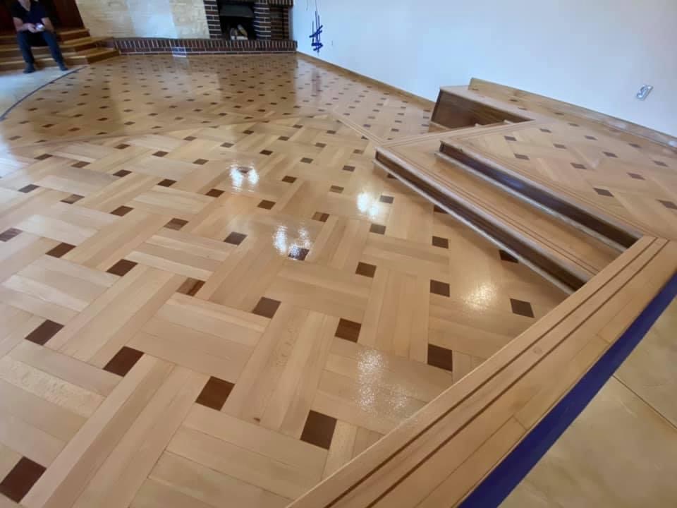 Parkiet ułożony w jodełkę z ciemnymi wstawkami, po renowacji, z widocznym fragmentem schodów i listwą przypodłogową zabezpieczoną taśmą malarską.