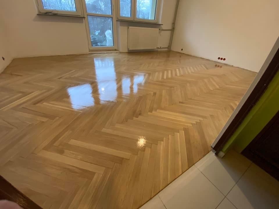 Odnowiony parkiet ułożony w jodełkę, pokryty błyszczącym lakierem, widok na okno z grzejnikiem i fragment ściany z wystającymi kablami elektrycznymi.