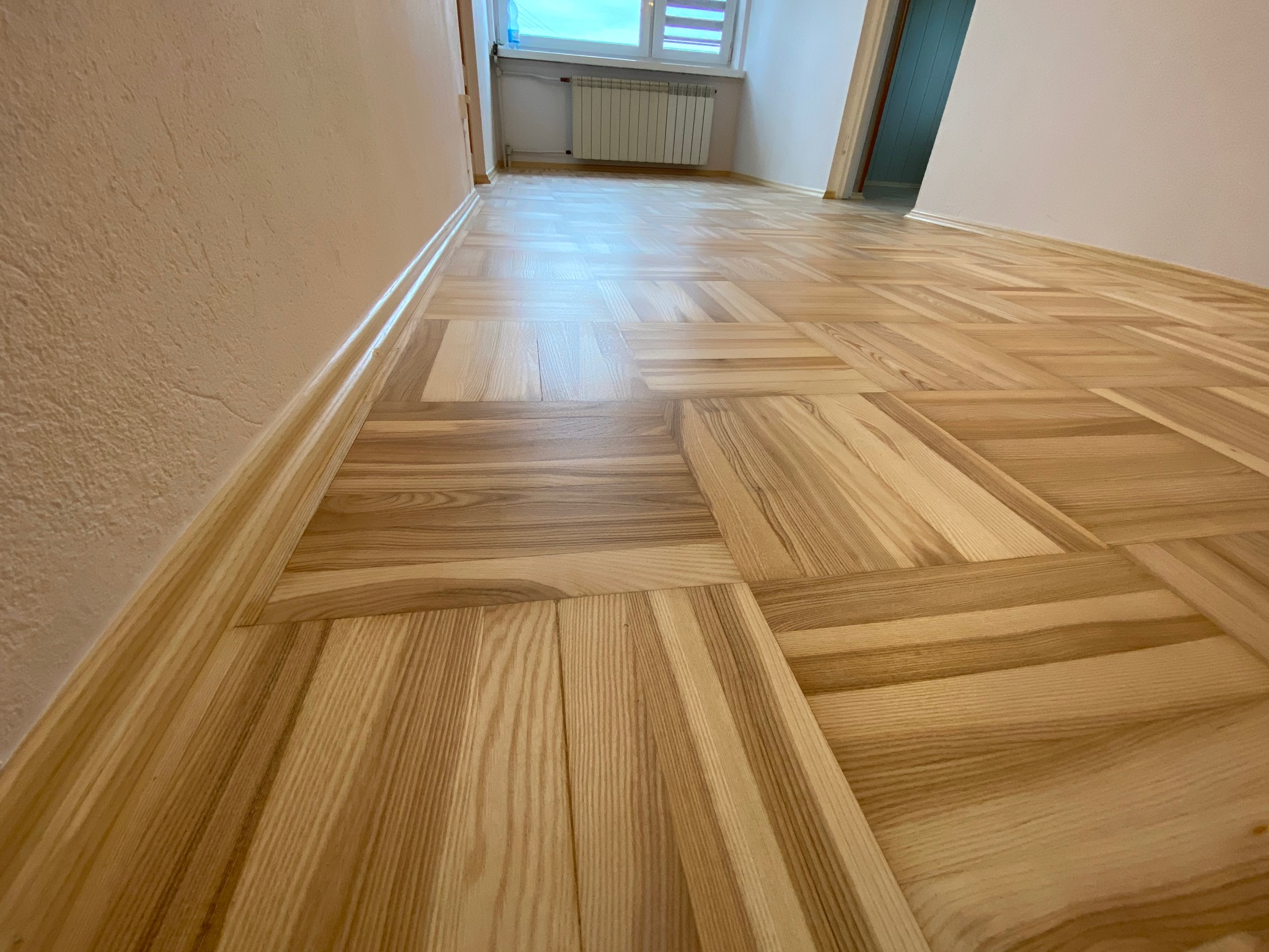 Parkiet ułożony w geometryczny wzór, świeżo po renowacji, z widocznymi słojami drewna i listwami przypodłogowymi przy ścianie w odcieniach beżu.