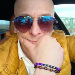 Autoportret taksówkarza w okularach przeciwsłonecznych z widocznym odbiciem wnętrza samochodu i mostu w szkle, ubrany w żółtą kurtkę, z bransoletkami na ręce.