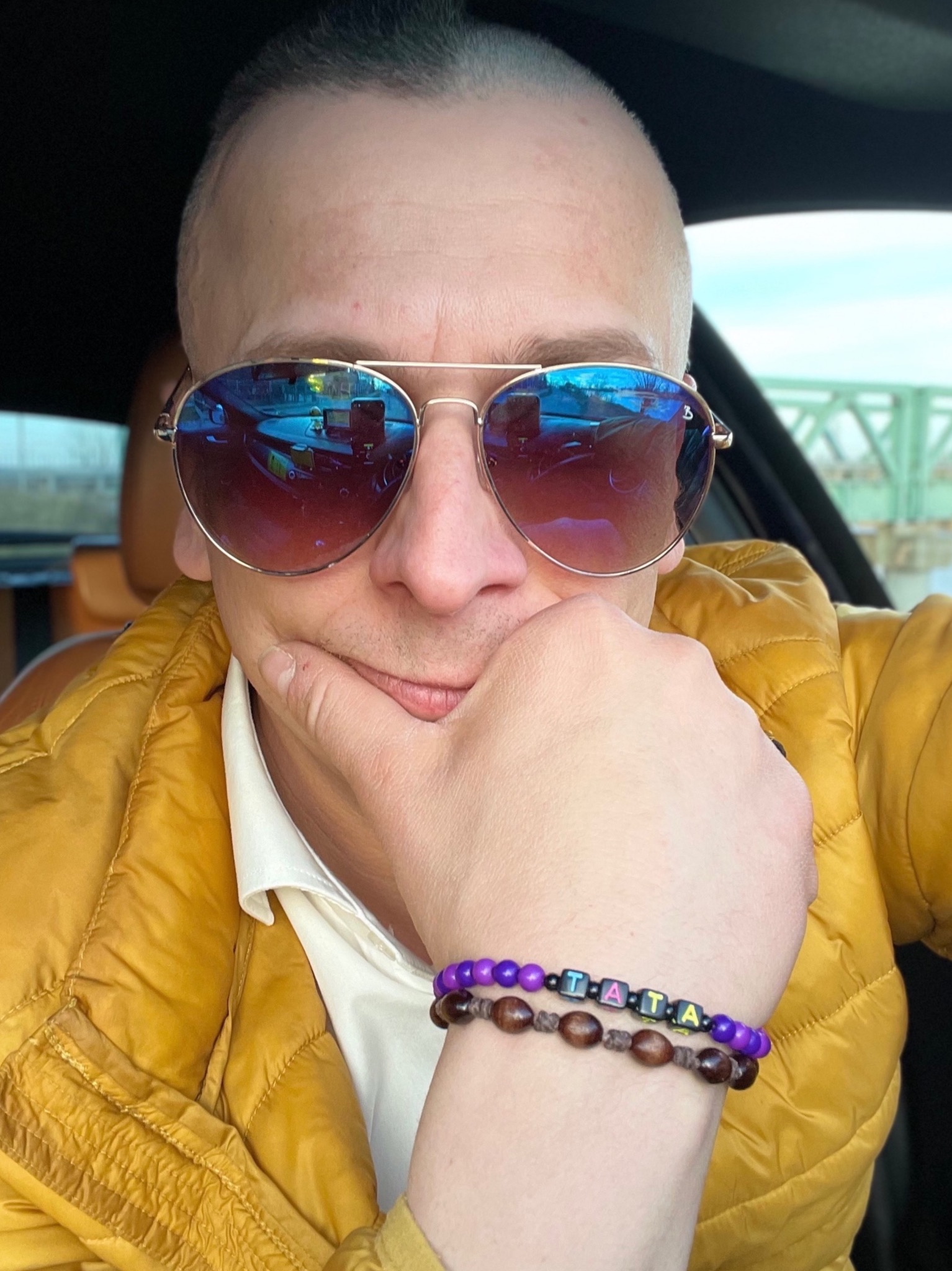 Autoportret taksówkarza w okularach przeciwsłonecznych z widocznym odbiciem wnętrza samochodu i mostu w szkle, ubrany w żółtą kurtkę, z bransoletkami na ręce.