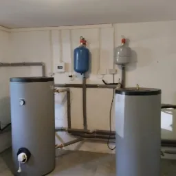 Dwa szare bojlery z czarnymi pokrywami, podłączone do szarych rur, z niebieskim i szarym zbiornikiem wyrównawczym na ścianie w tle.