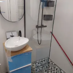 Nowoczesna łazienka z umywalką nablatową w kształcie misy, szafką w niebieskim kolorze zabezpieczoną folią ochronną, kabiną prysznicową ze szklaną ścianą, czarną armaturą i wzorzystą podłogą...