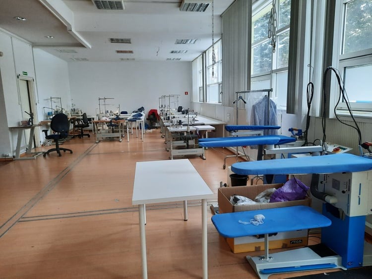 Przestronna szwalnia z wieloma stanowiskami pracy wyposażonymi w maszyny do szycia, stoły do krojenia i prasowania, widok na duże okna i parkiet w tle.