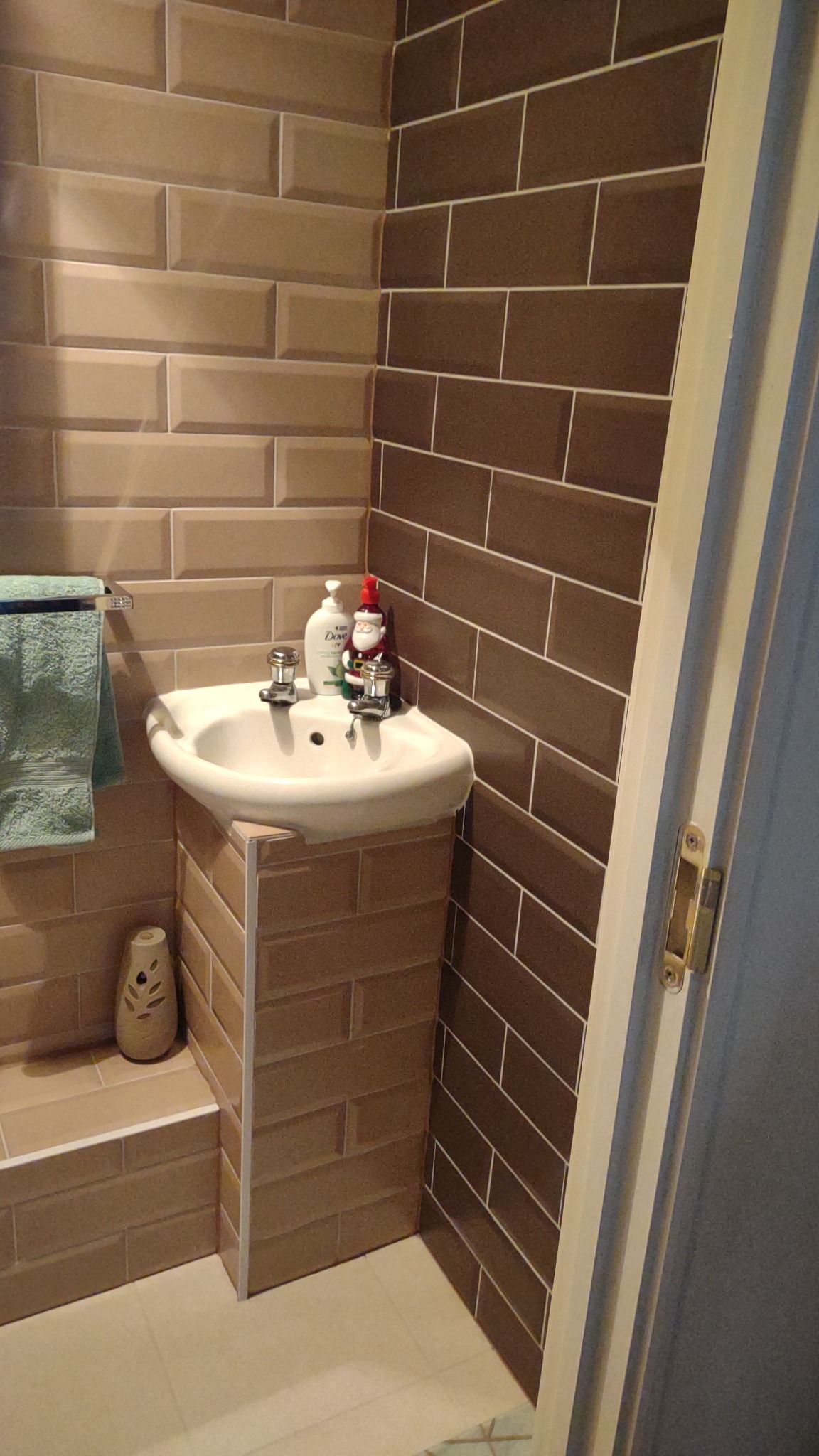 Umywalka narożna z postumentem obłożona brązowymi płytkami imitującymi cegłę, ściany wyłożone podobnymi płytkami w dwóch odcieniach brązu, na umywalce mydło w płynie i figurka Mikołaja.