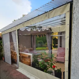 Zadaszona altana z transparentnymi ścianami bocznymi, meblami z palet i roślinami w donicach, widok z zewnątrz. Lekka konstrukcja z przezroczystym dachem.