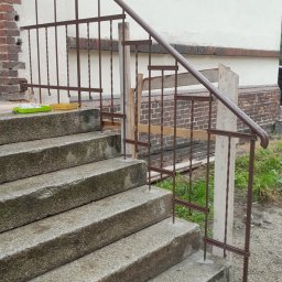 PPHU ROB-MAR Robert Marek - Montaż brązowej, metalowej balustrady schodowej zewnętrznej z drewnianym wspornikiem, na betonowych schodach przed budynkiem z ceglanym fragmentem elewacji.