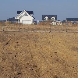 PPHU ROB-MAR Robert Marek - Nowoczesne ogrodzenie panelowe na tle nowo wybudowanych domów jednorodzinnych. Widok na brązową ziemię i czarne słupki ogrodzeniowe.