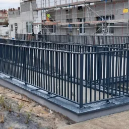 Nowoczesna, prosta balustrada z ciemnego metalu zabezpieczająca teren przed budynkiem w trakcie budowy, widoczni robotnicy na rusztowaniu.