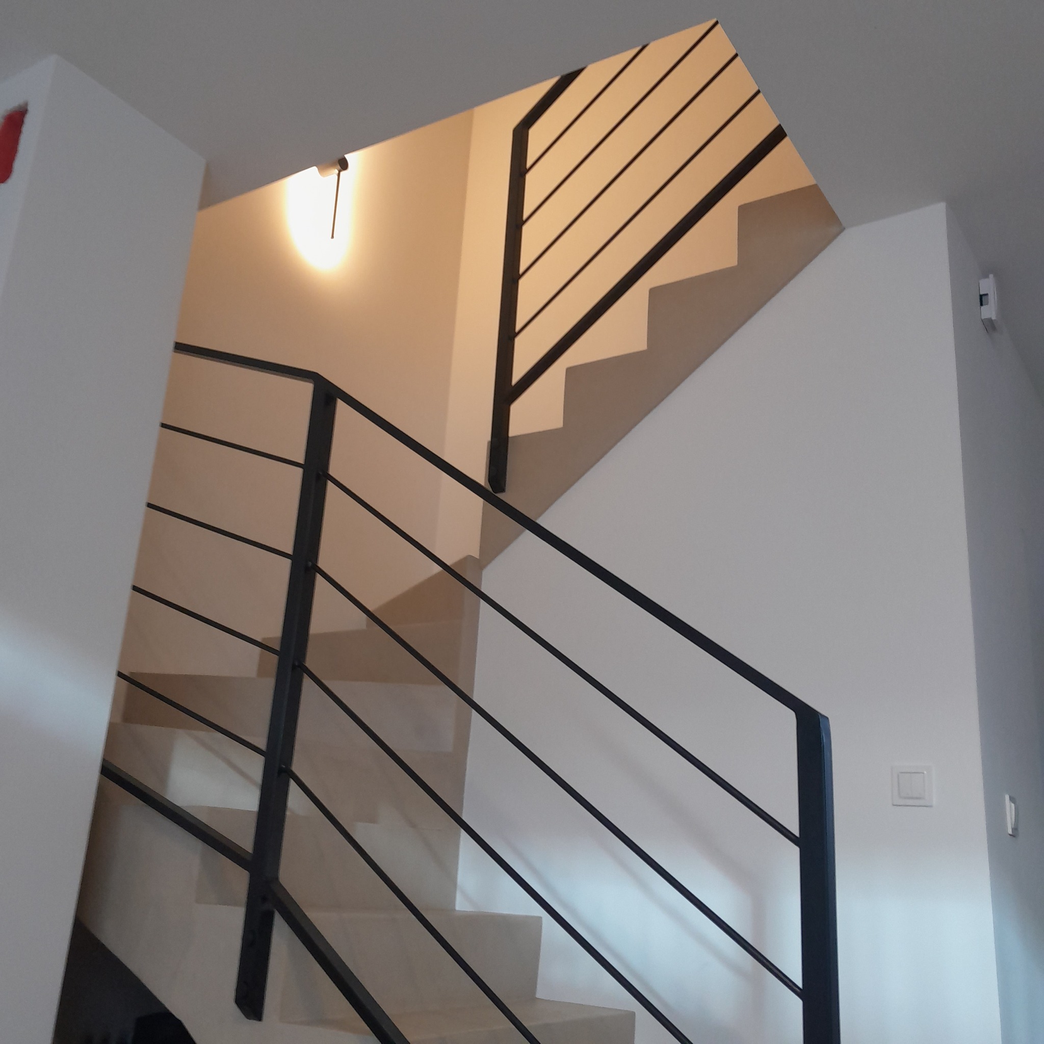 Nowoczesna balustrada schodowa z czarnych, cienkich prętów, widoczna z dołu, oświetlona ciepłym światłem, z białymi ścianami i schodami.