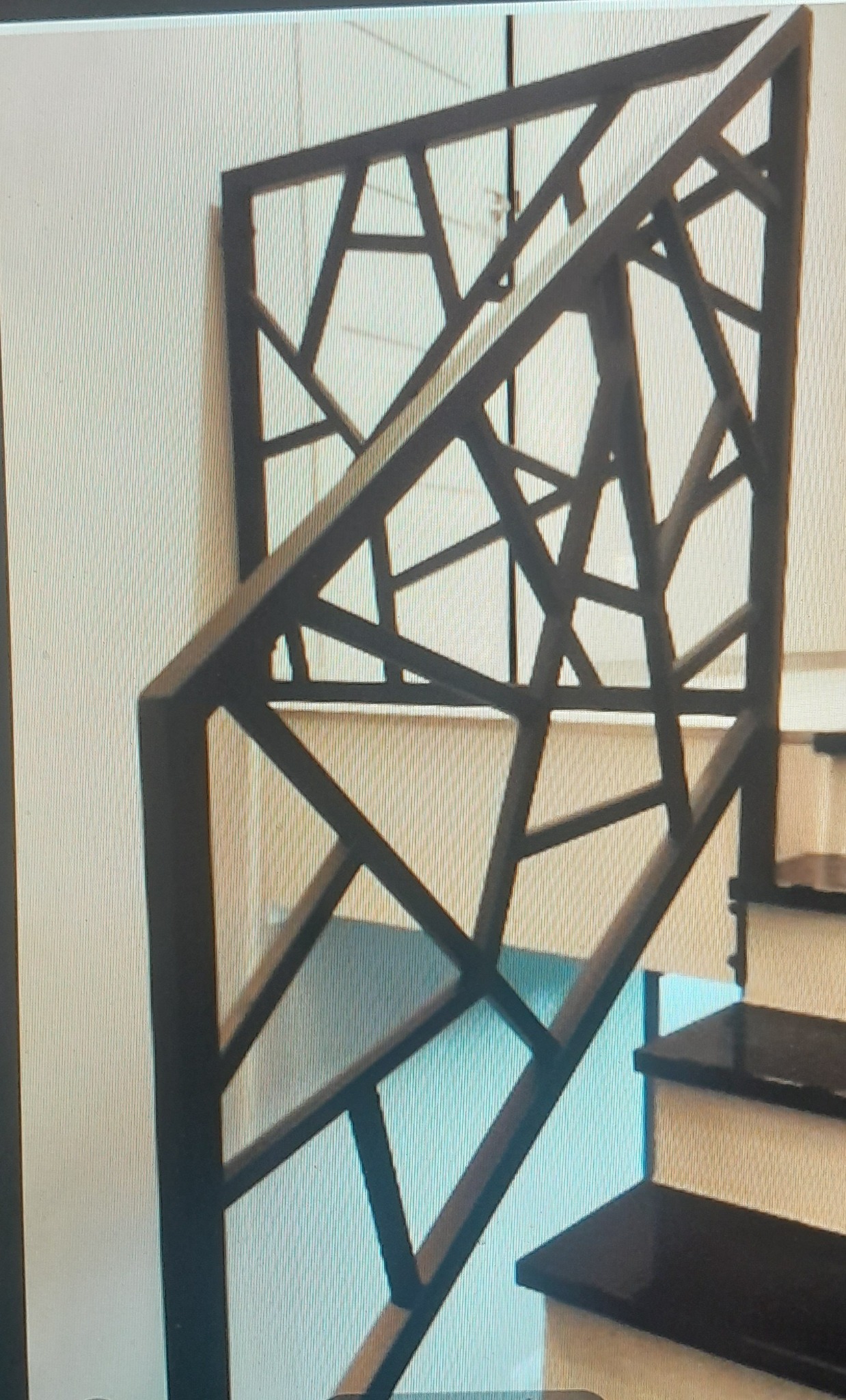 Nowoczesna balustrada schodowa z metalu o geometrycznym wzorze, pomalowana na ciemny kolor, widoczna z bliska, z fragmentem schodów w tle.