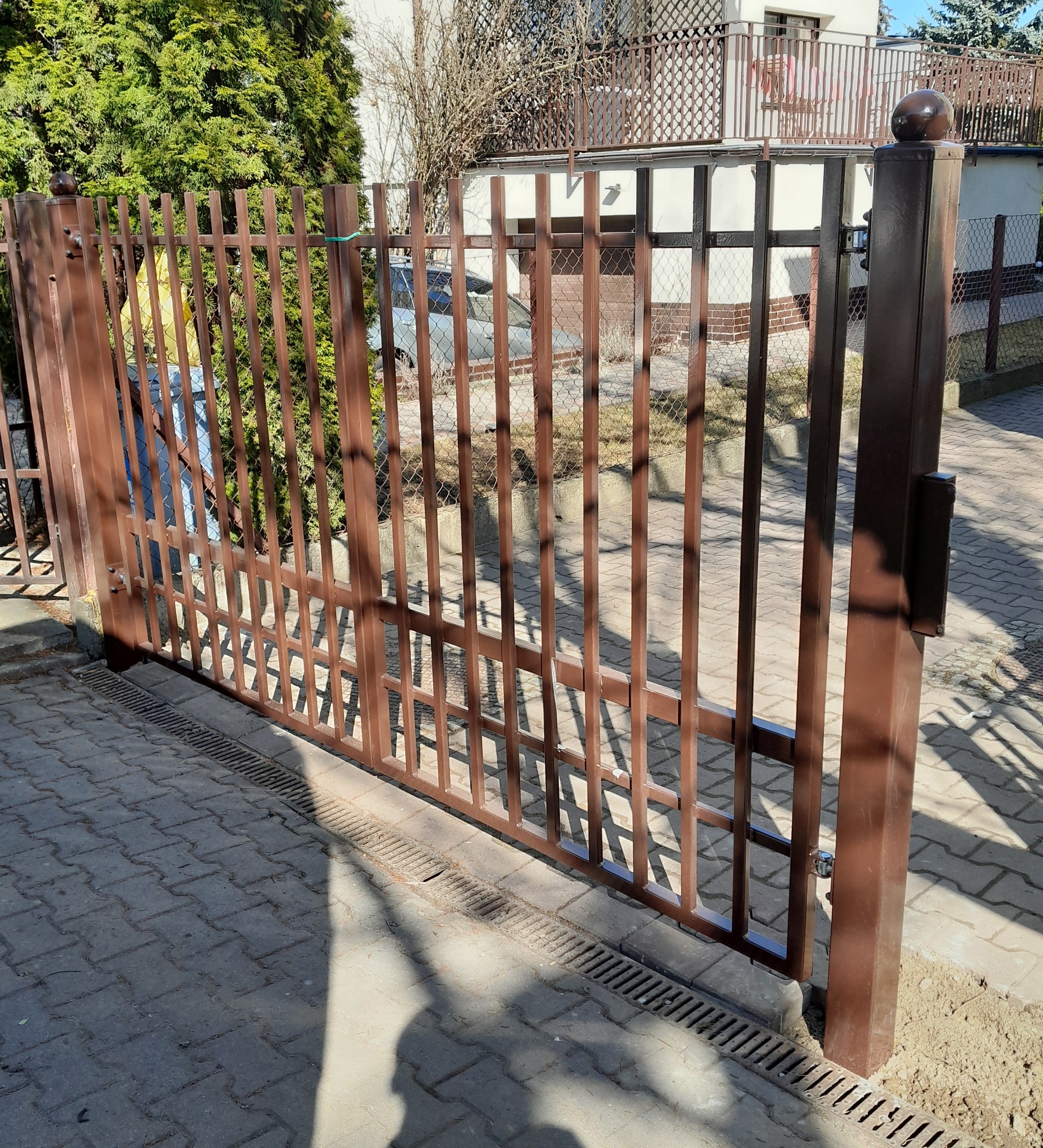 Brązowa, metalowa brama wjazdowa z pionowymi prętami, widoczny mechanizm zamka, kostka brukowa i trawnik w tle, w słoneczny dzień.