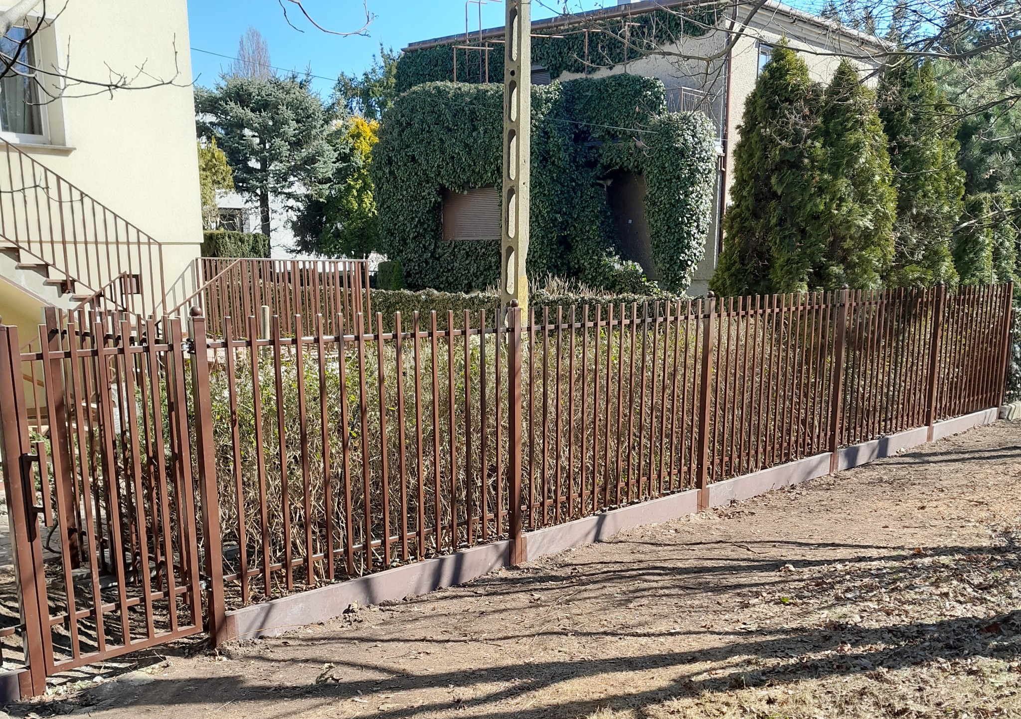 Brązowe metalowe ogrodzenie z furtką, widok na dom z zielonym bluszczem i drzewa w tle, słoneczny dzień.