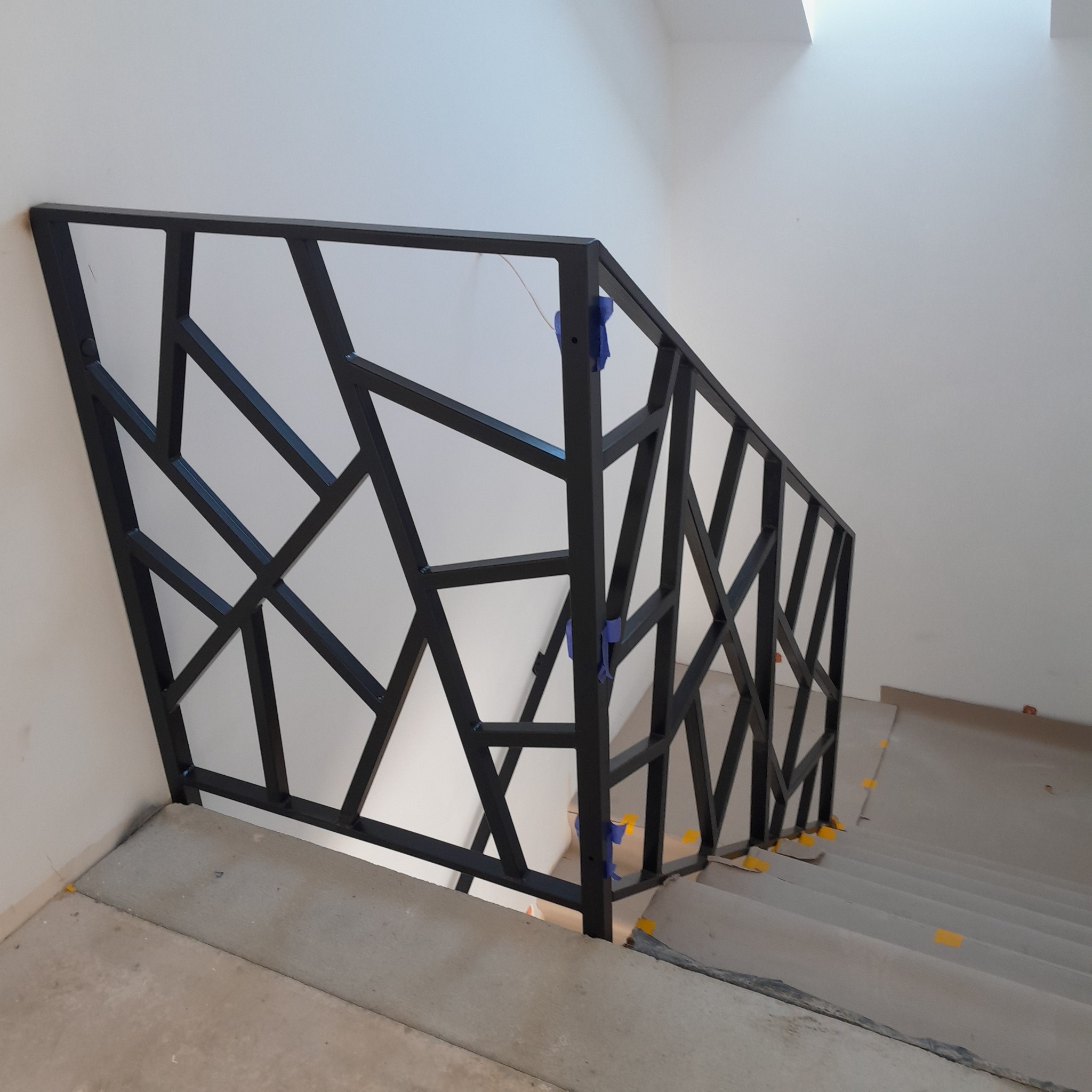 Czarna, metalowa balustrada schodowa o geometrycznym wzorze, zabezpieczona niebieską folią, zamontowana na surowych, betonowych schodach w jasnym wnętrzu.