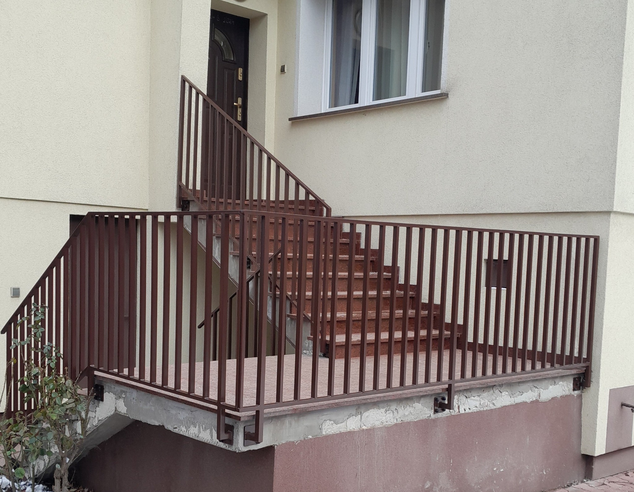 Brązowa balustrada schodowa z pionowymi szczeblami, zabezpieczająca wejście do budynku z jasną elewacją i brązowymi drzwiami.
