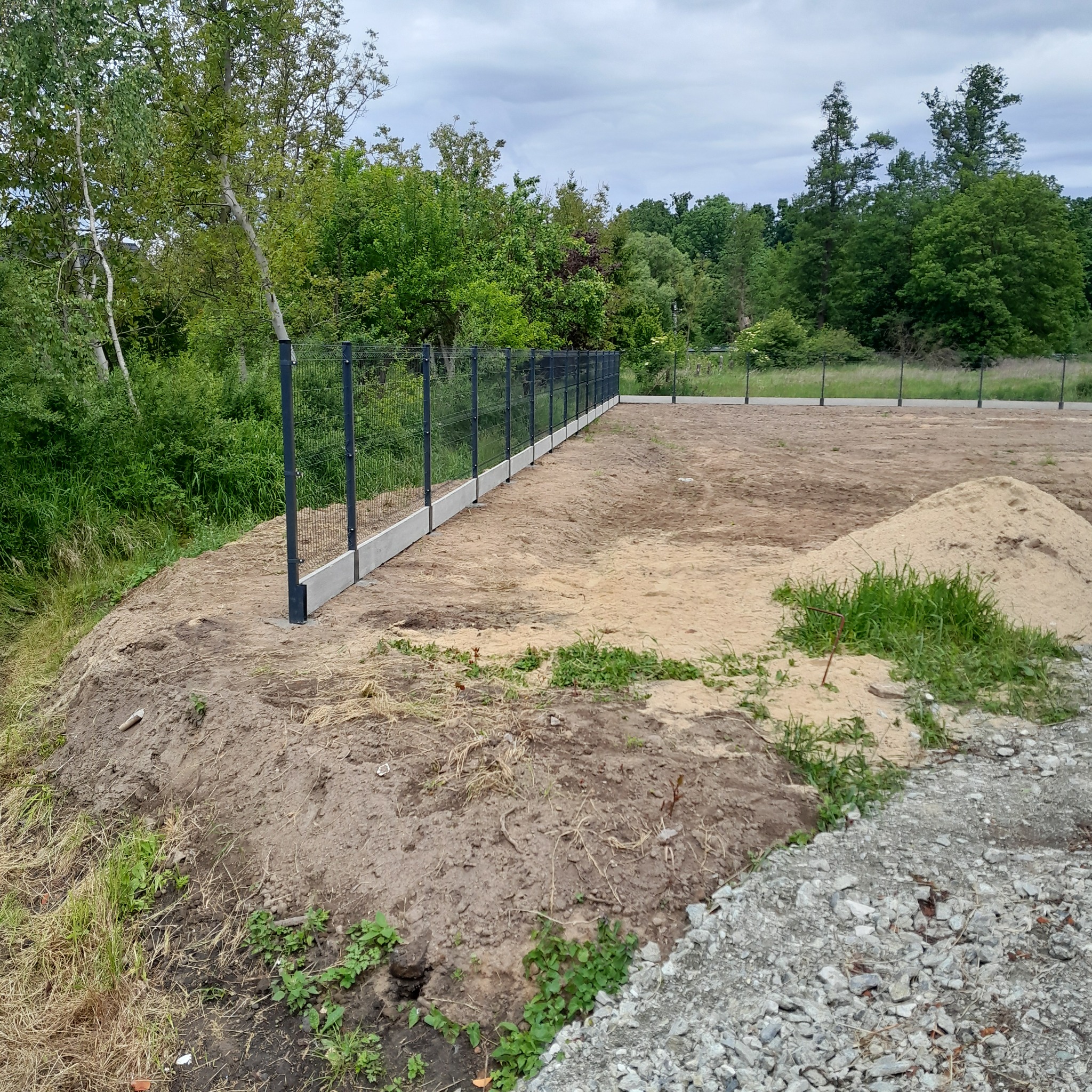 Nowo postawione, panelowe ogrodzenie z podmurówką betonową, widoczne na tle zieleni drzew i nieba. Na pierwszym planie ziemia i usypany piasek.