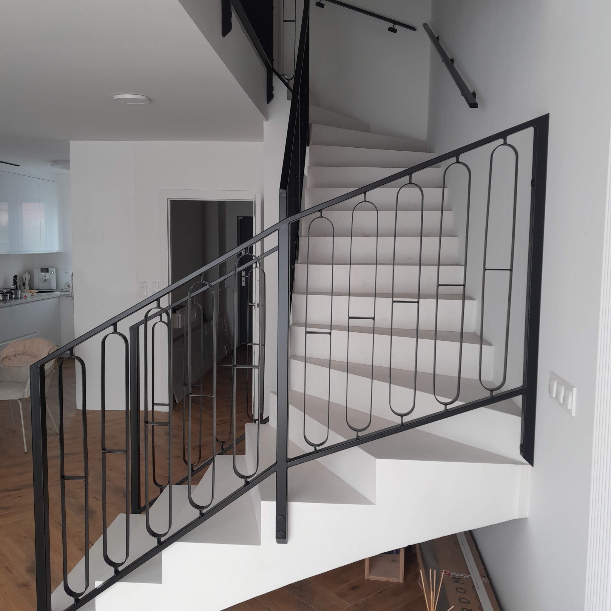 Białe schody z czarną, metalową balustradą o nowoczesnym wzorze, w jasnym wnętrzu domu. Widoczny fragment kuchni i drzwi. Minimalistyczny design.