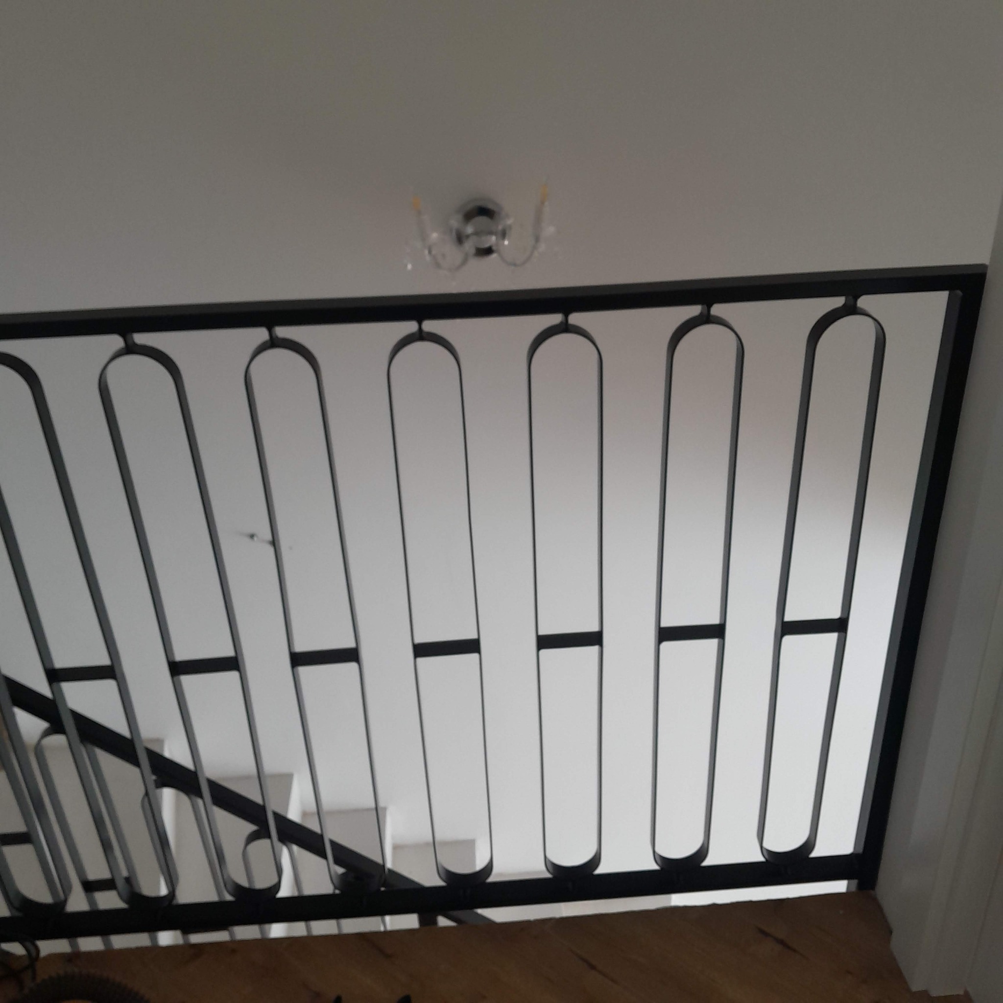Nowoczesna, czarna balustrada schodowa z metalowymi prętami o zaokrąglonych końcach, widok od dołu na tle białego sufitu z lampą.