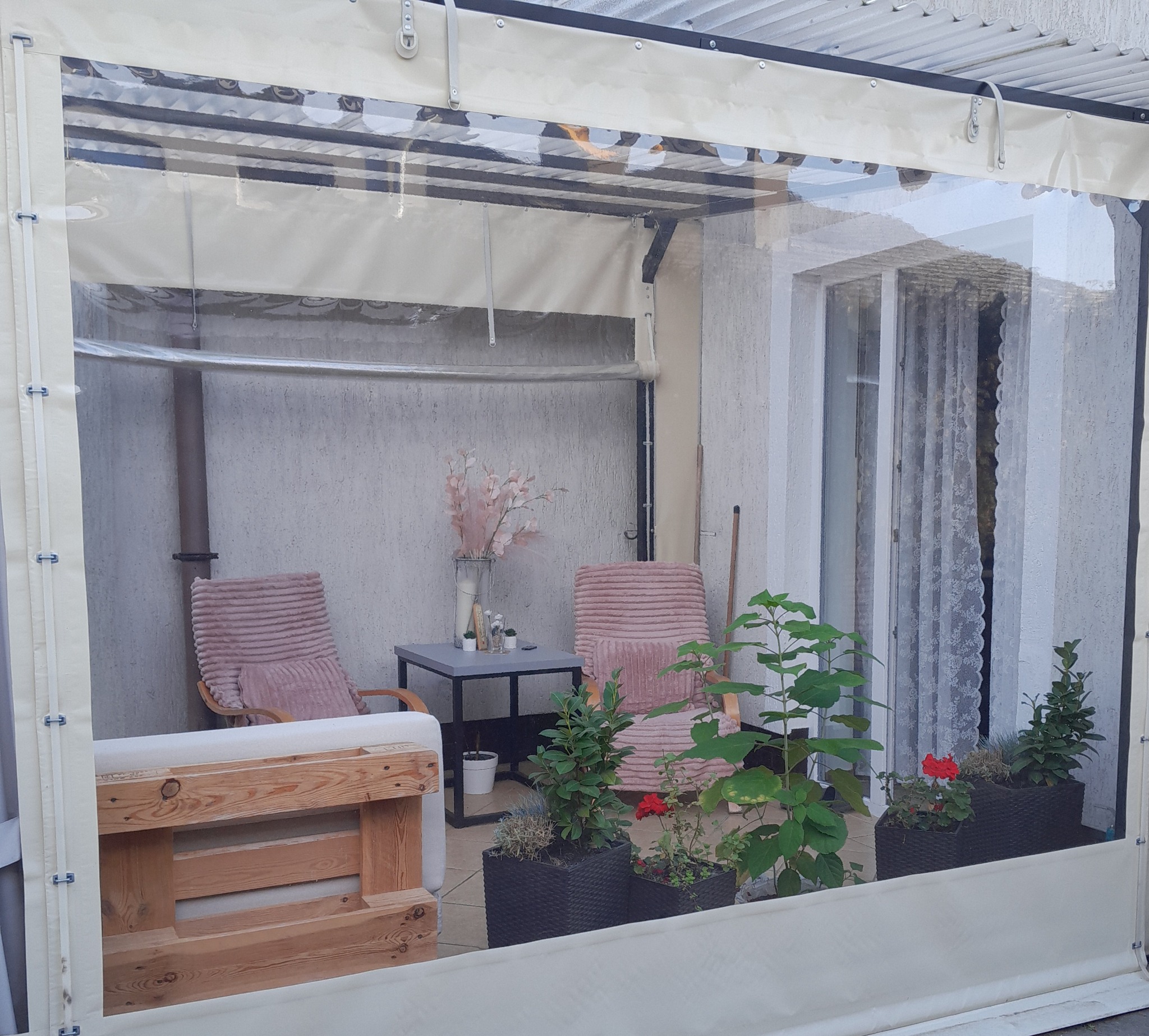 Zadaszony taras z przezroczystymi ścianami, meblami ogrodowymi i roślinami w donicach. Widoczne krzesła, stolik i drewniana konstrukcja. Oświetlenie naturalne.