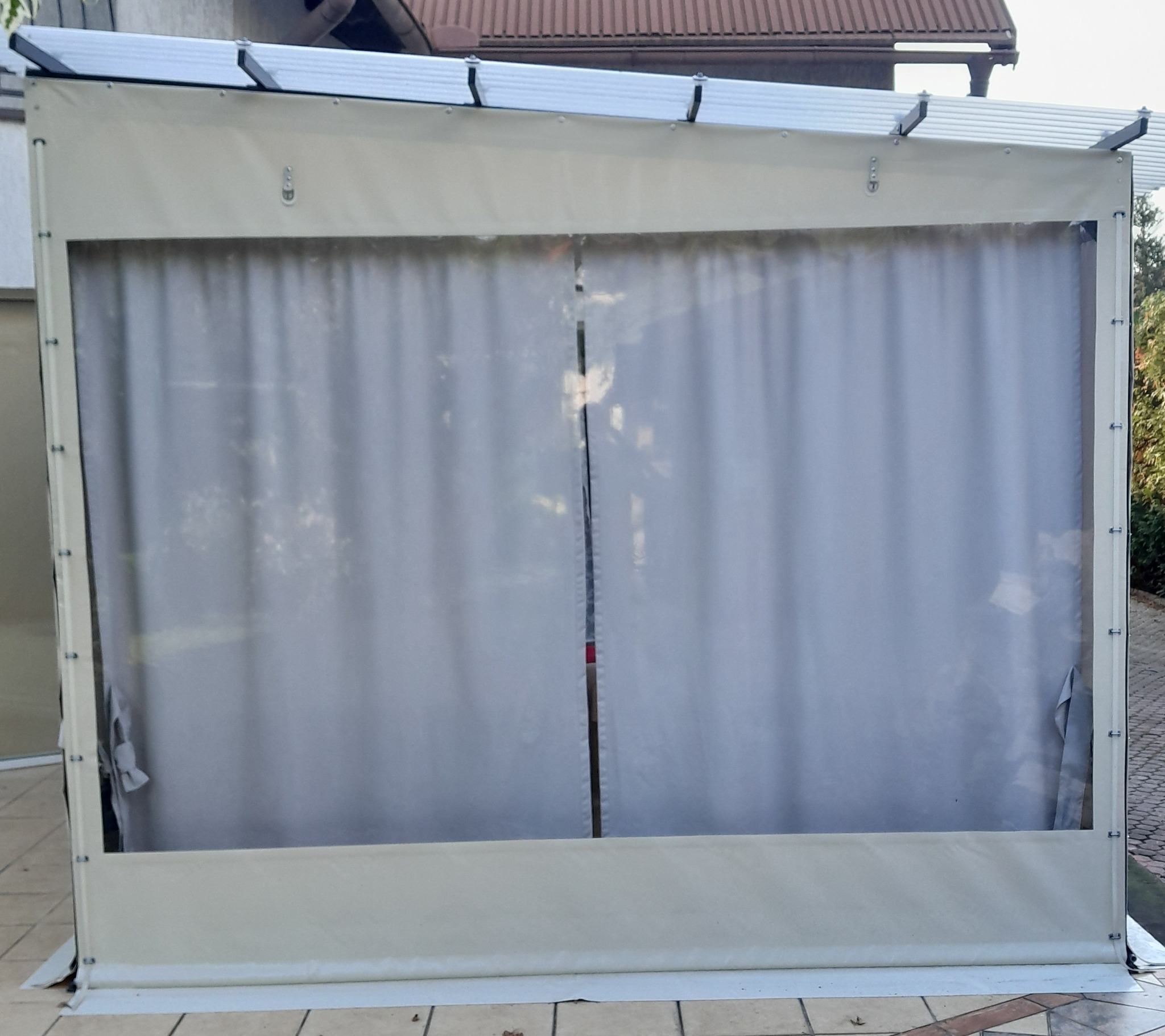 Beżowa konstrukcja z zasłonami, prawdopodobnie osłona tarasu lub werandy, z widocznymi detalami montażu i elementami dachu, na tle budynku.