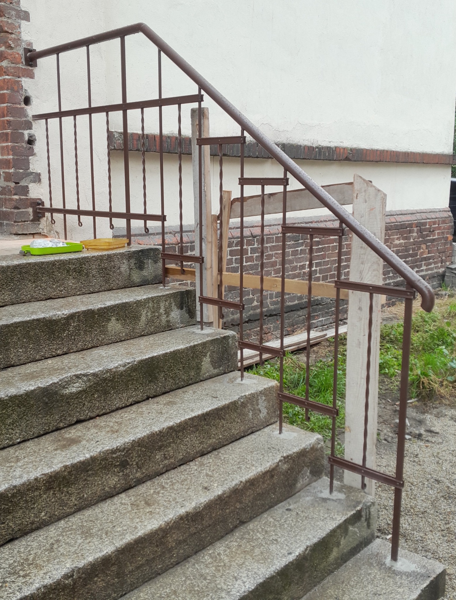 Montaż brązowej, metalowej balustrady schodowej zewnętrznej z drewnianym wspornikiem, na betonowych schodach przed budynkiem z ceglanym fragmentem elewacji.