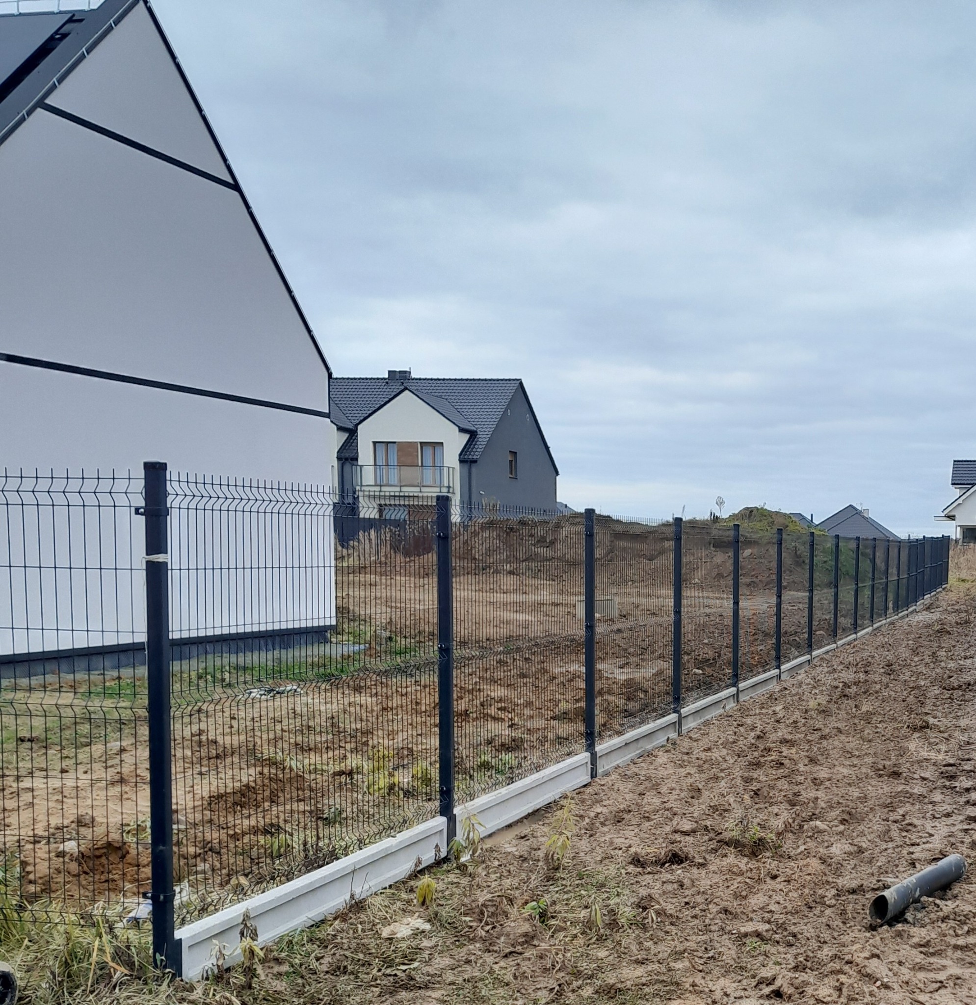 Nowoczesne ogrodzenie panelowe z podmurówką betonową, widok z boku na tle domów jednorodzinnych w pochmurny dzień. Słupki w kolorze grafitowym.