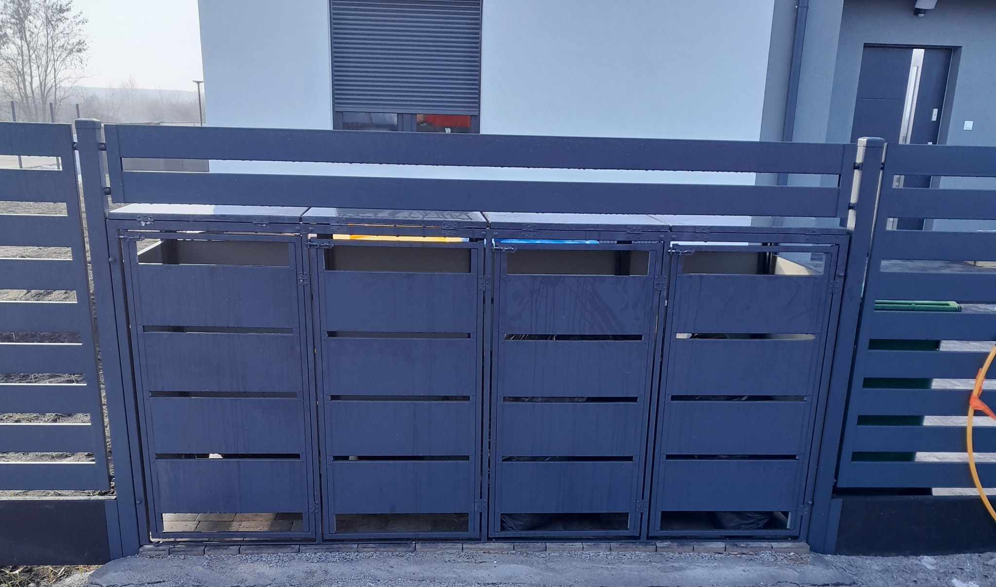 Nowoczesne, metalowe ogrodzenie zintegrowane z zabudową śmietnikową, pomalowane na grafitowy kolor, z poziomymi szczeblami, na tle jasnego budynku.