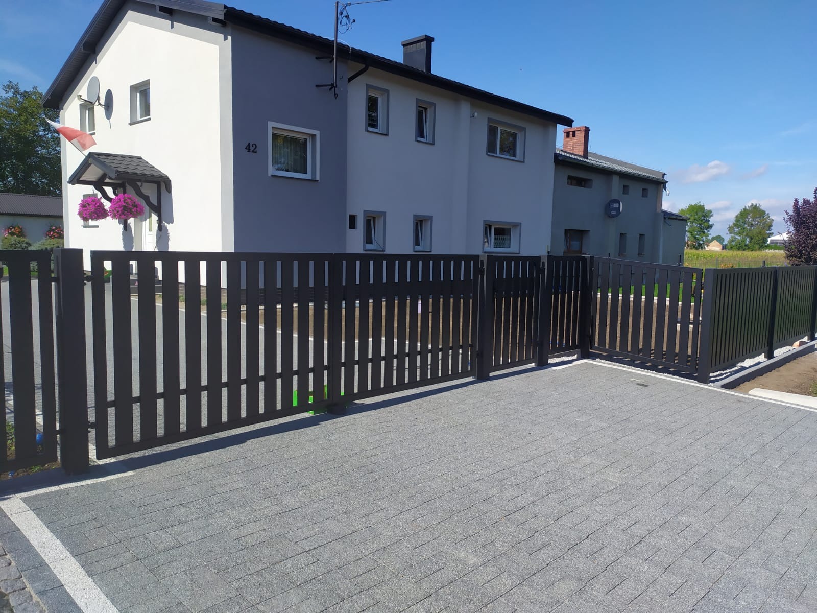 Nowoczesne, grafitowe ogrodzenie posesji z furtką, na tle dwukondygnacyjnego domu z numerem 42, w słoneczny dzień. Kostka brukowa na podjeździe.