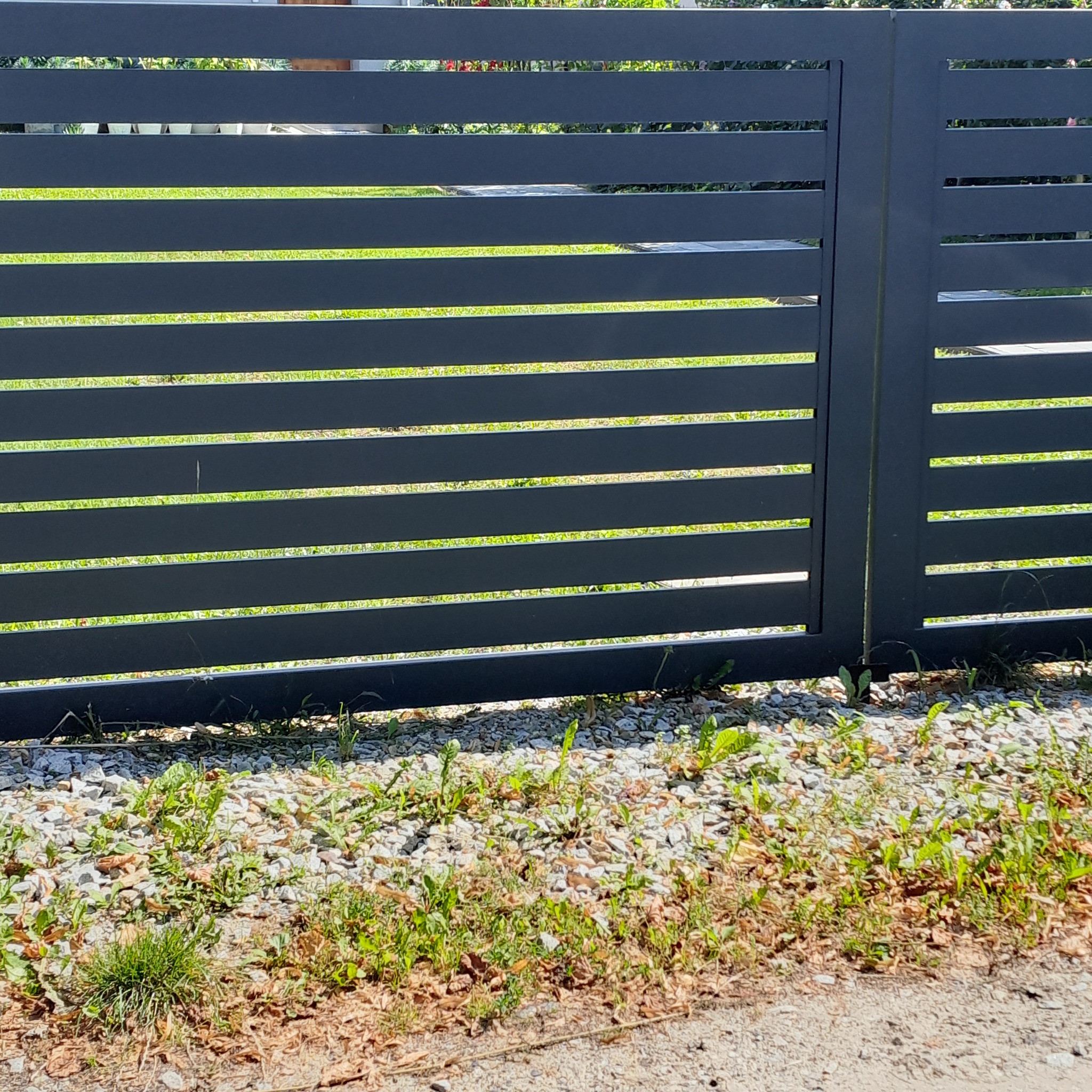 Nowoczesne, poziome ogrodzenie panelowe w kolorze grafitowym, z widocznymi prześwitami na tle zieleni. Minimalistyczny design, kamieniste podłoże.