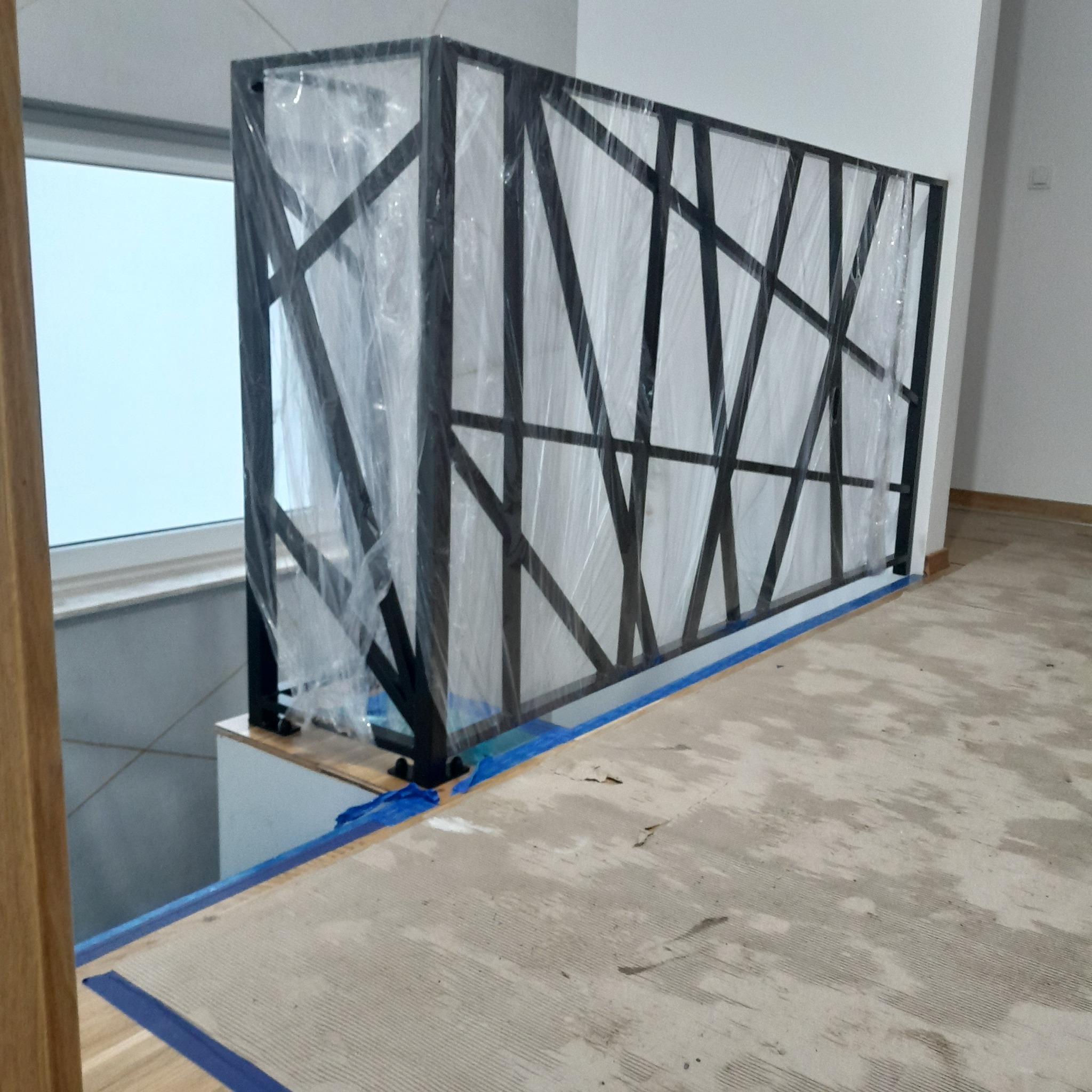 Nowoczesna balustrada metalowa z geometrycznym wzorem, zabezpieczona folią ochronną podczas prac wykończeniowych. Widoczne surowe podłoże i okno w tle.