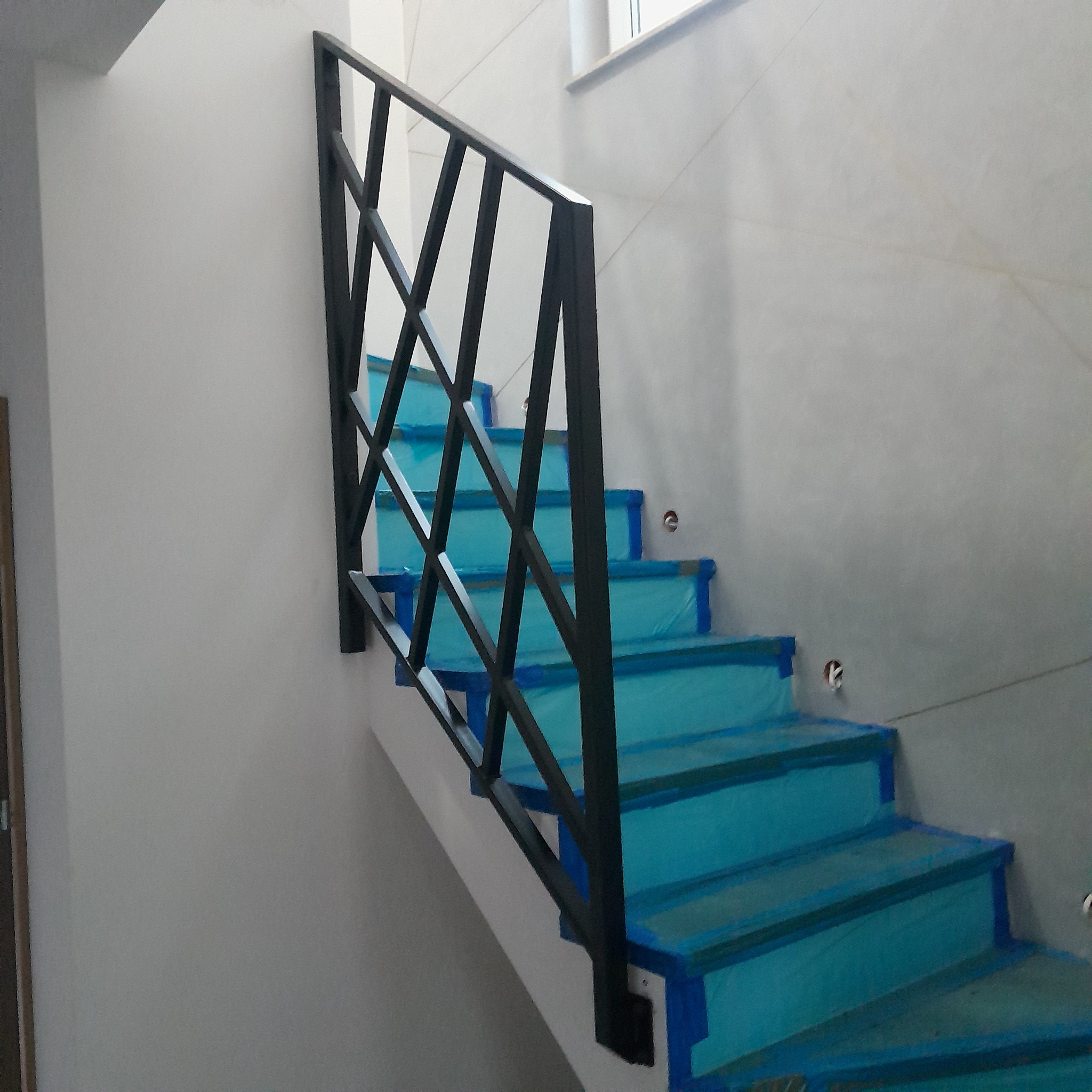 Nowoczesne schody z czarną, metalową balustradą o geometrycznym wzorze, zabezpieczone folią ochronną podczas prac wykończeniowych w jasnym wnętrzu.