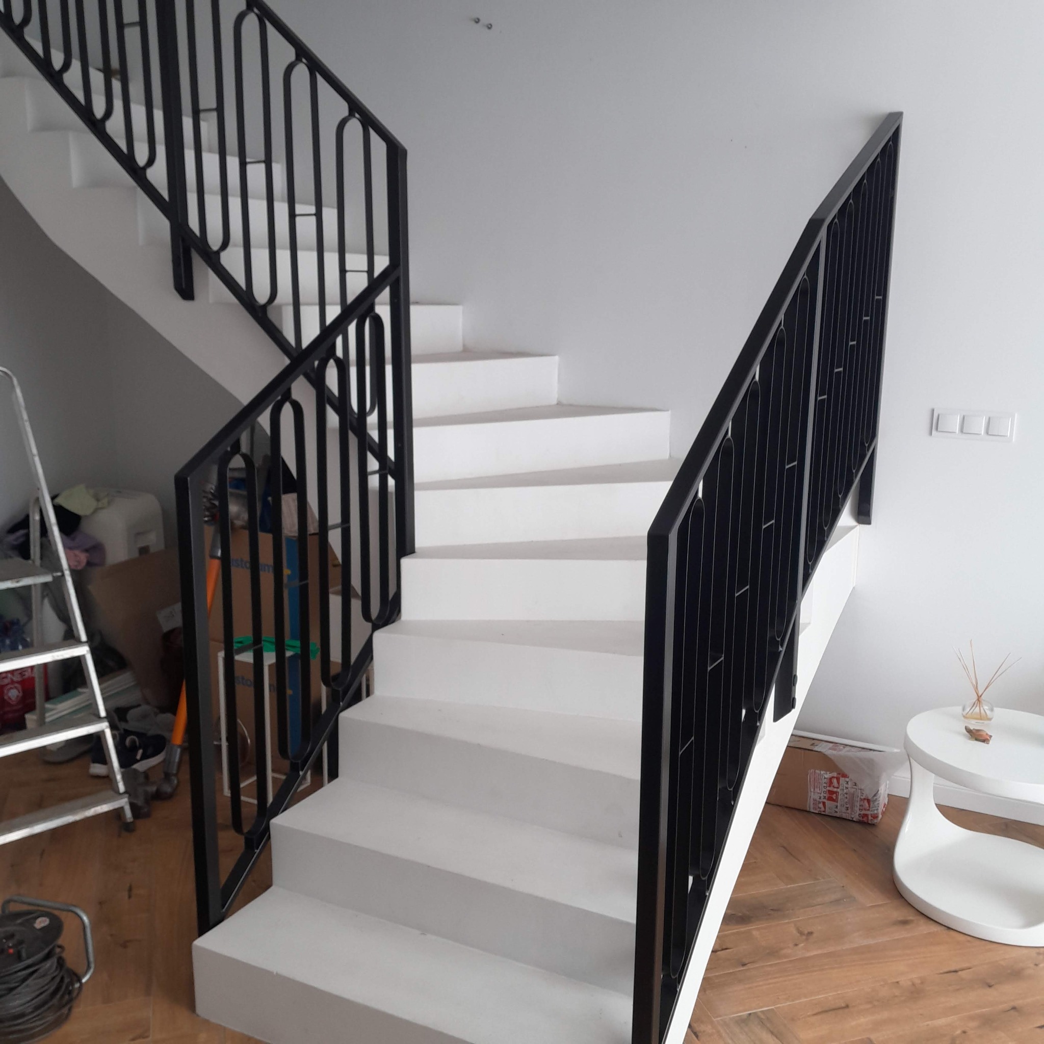 Nowoczesne schody z białą konstrukcją i czarną, metalową balustradą o minimalistycznym designie. Widoczne detale wykończenia i jasne wnętrze.