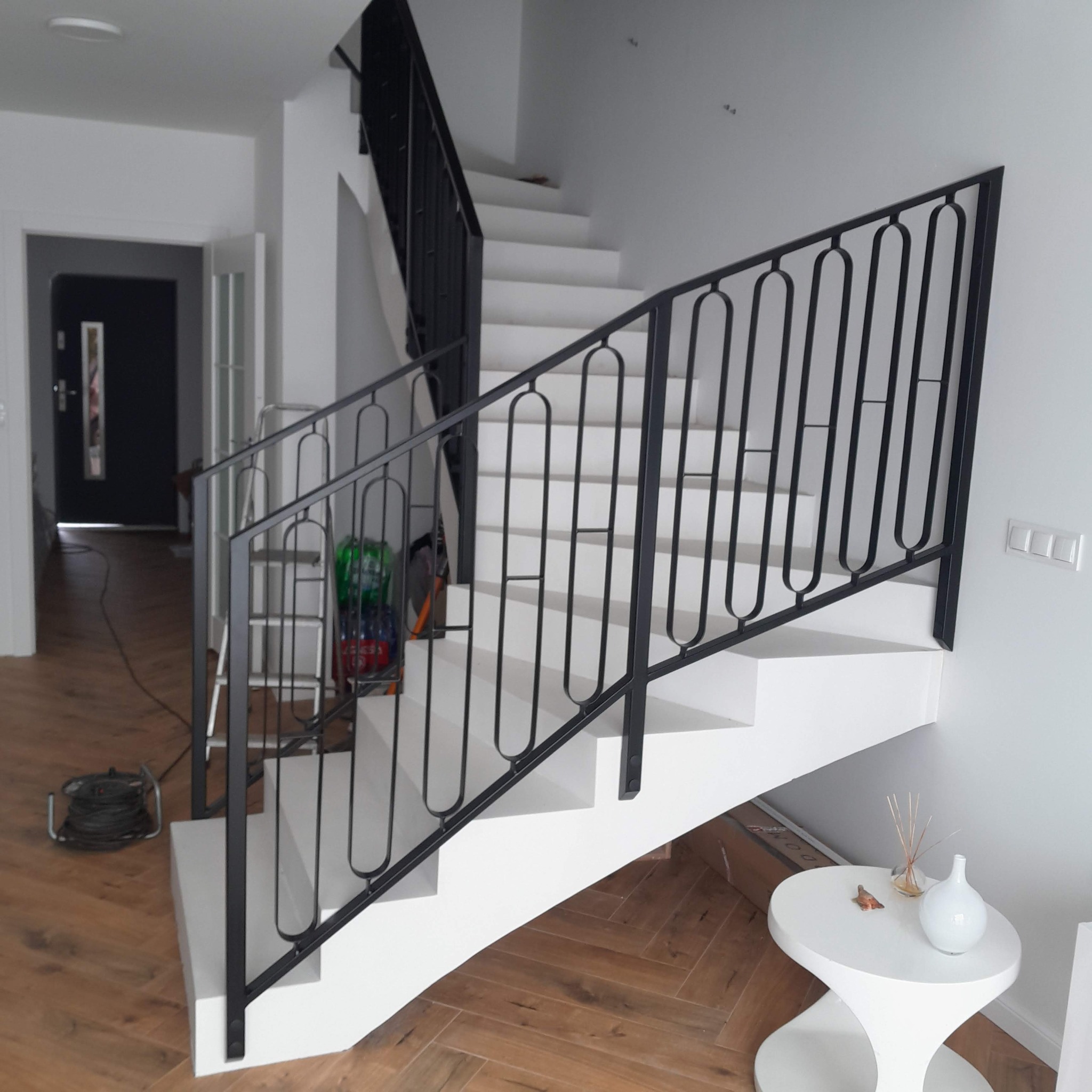 Nowoczesne, białe schody z czarną, metalową balustradą o dekoracyjnym wzorze. Widoczny fragment salonu z drewnianą podłogą i białym stolikiem.