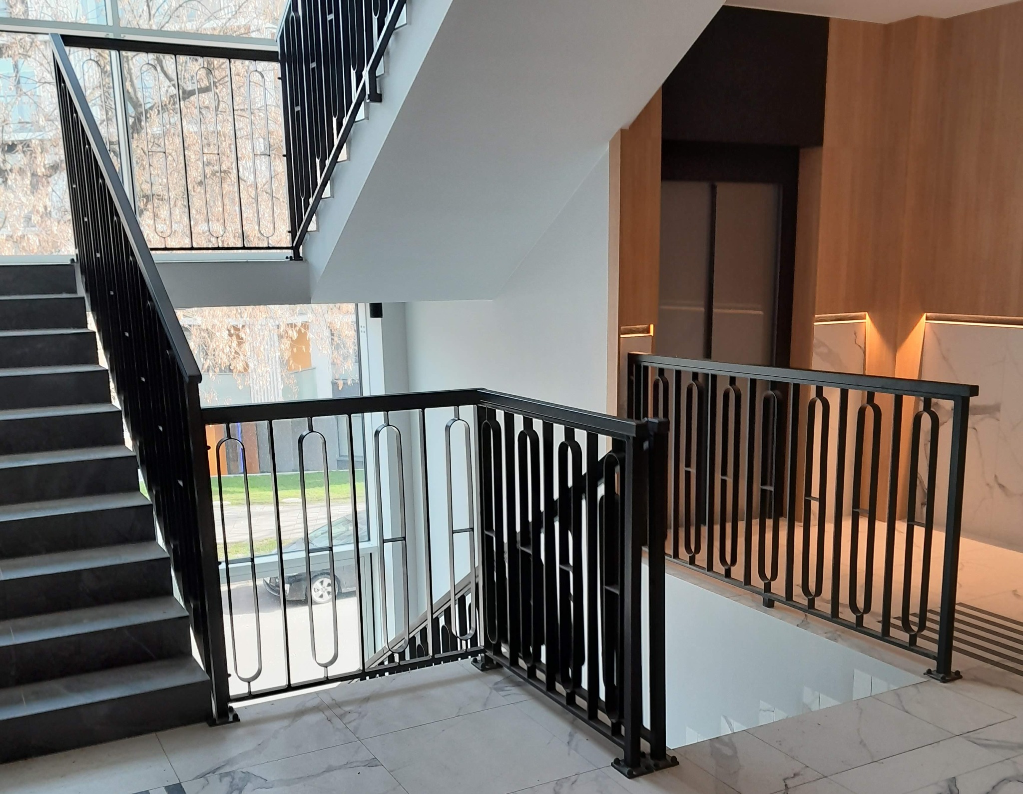 Nowoczesne schody z czarną metalową balustradą o unikalnym wzorze, widok z góry na marmurowe stopnie i jasne wnętrze budynku.