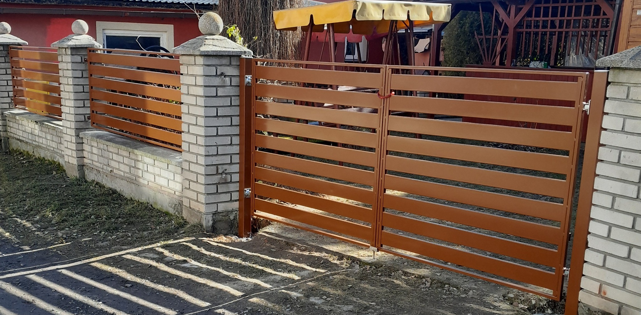 Nowoczesne ogrodzenie posesji z poziomych, brązowych paneli metalowych, osadzone na niskim murku z cegły. Brama dwuskrzydłowa zamykana na zasuwkę.