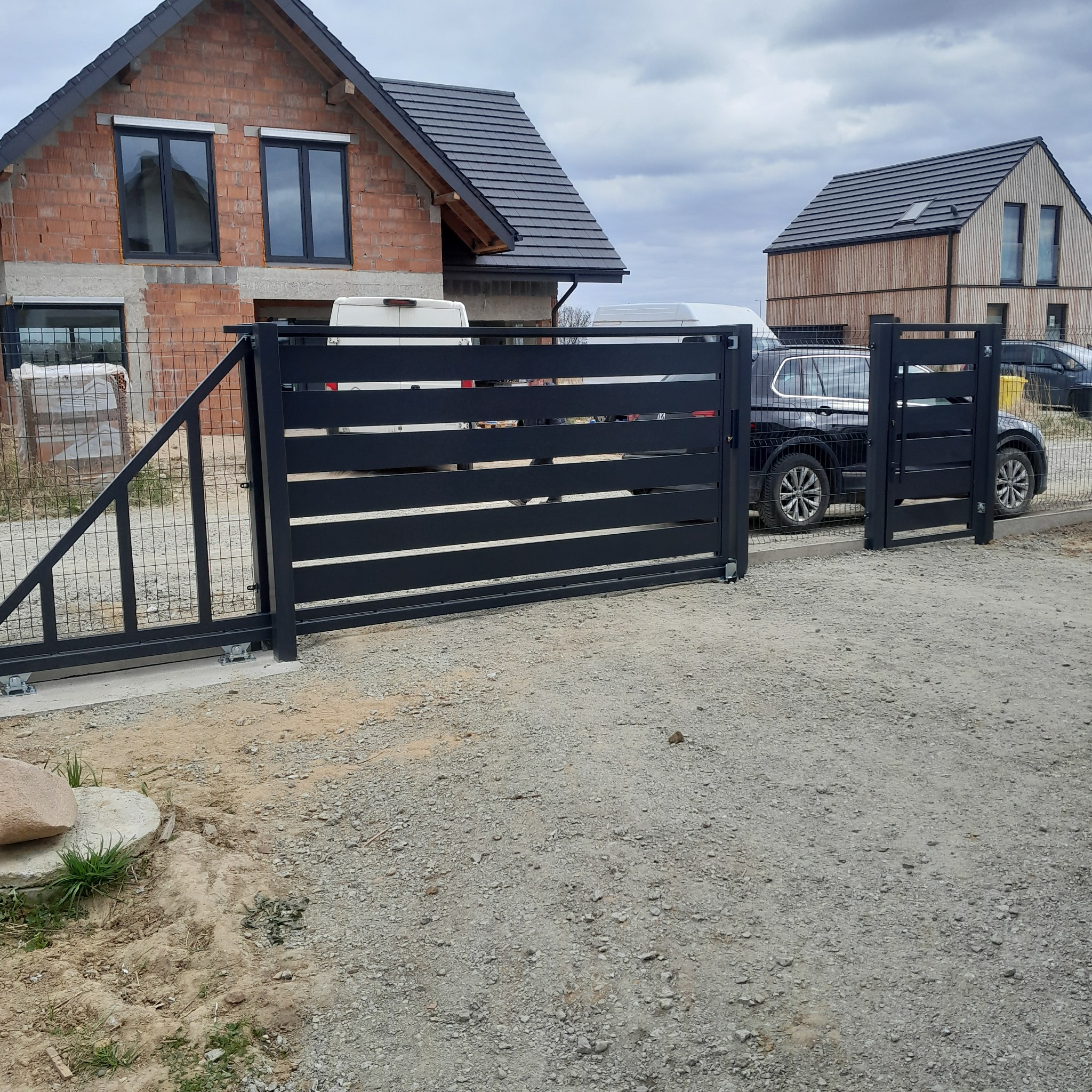 Nowoczesna, przesuwna brama wjazdowa z poziomymi panelami, zamontowana na posesji z domami w trakcie budowy. Widoczna siatka ogrodzeniowa i samochód osobowy.