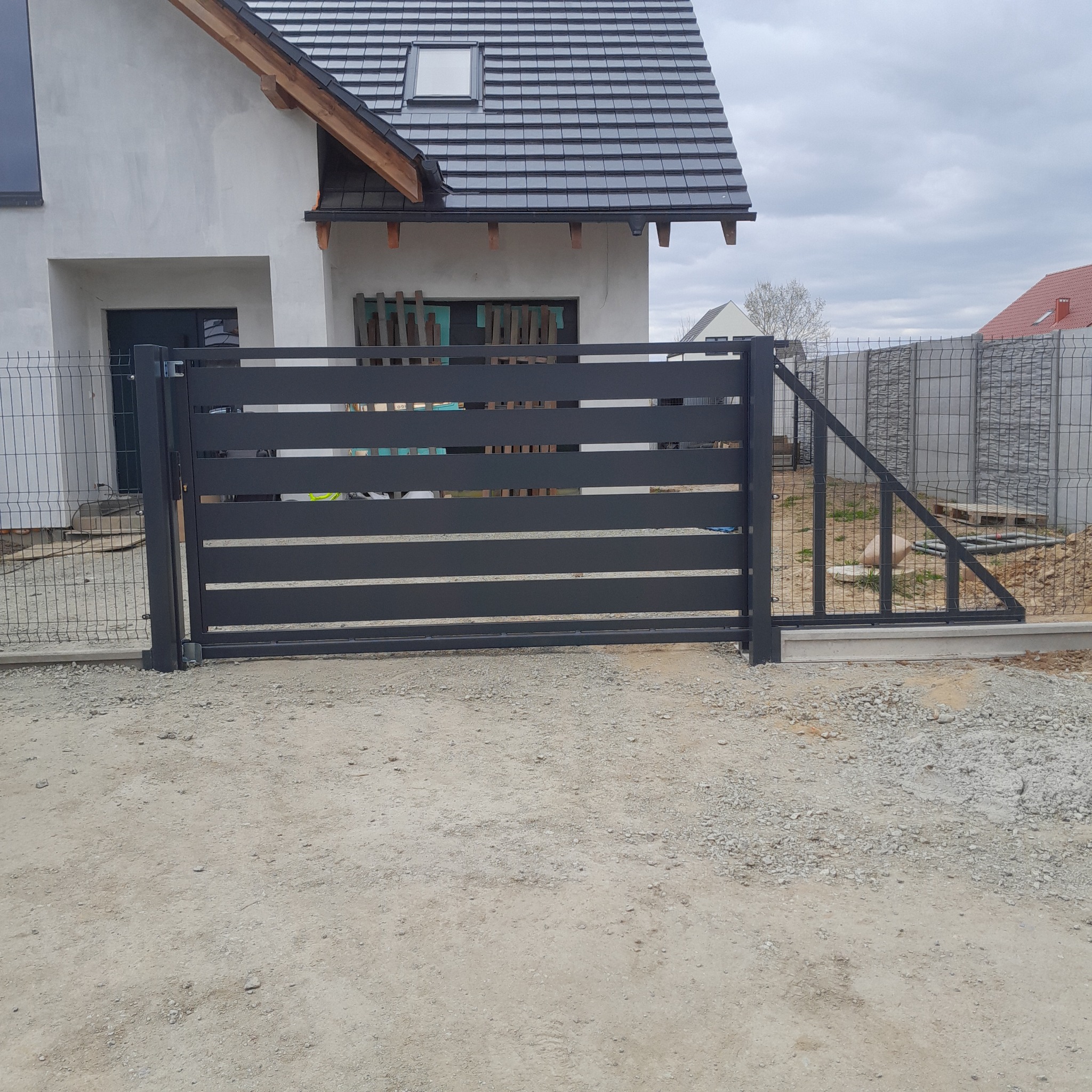 Nowoczesna, pozioma brama wjazdowa z metalowymi panelami w kolorze antracytowym, zamontowana przy domu z szarą elewacją i ogrodzeniem z siatki.