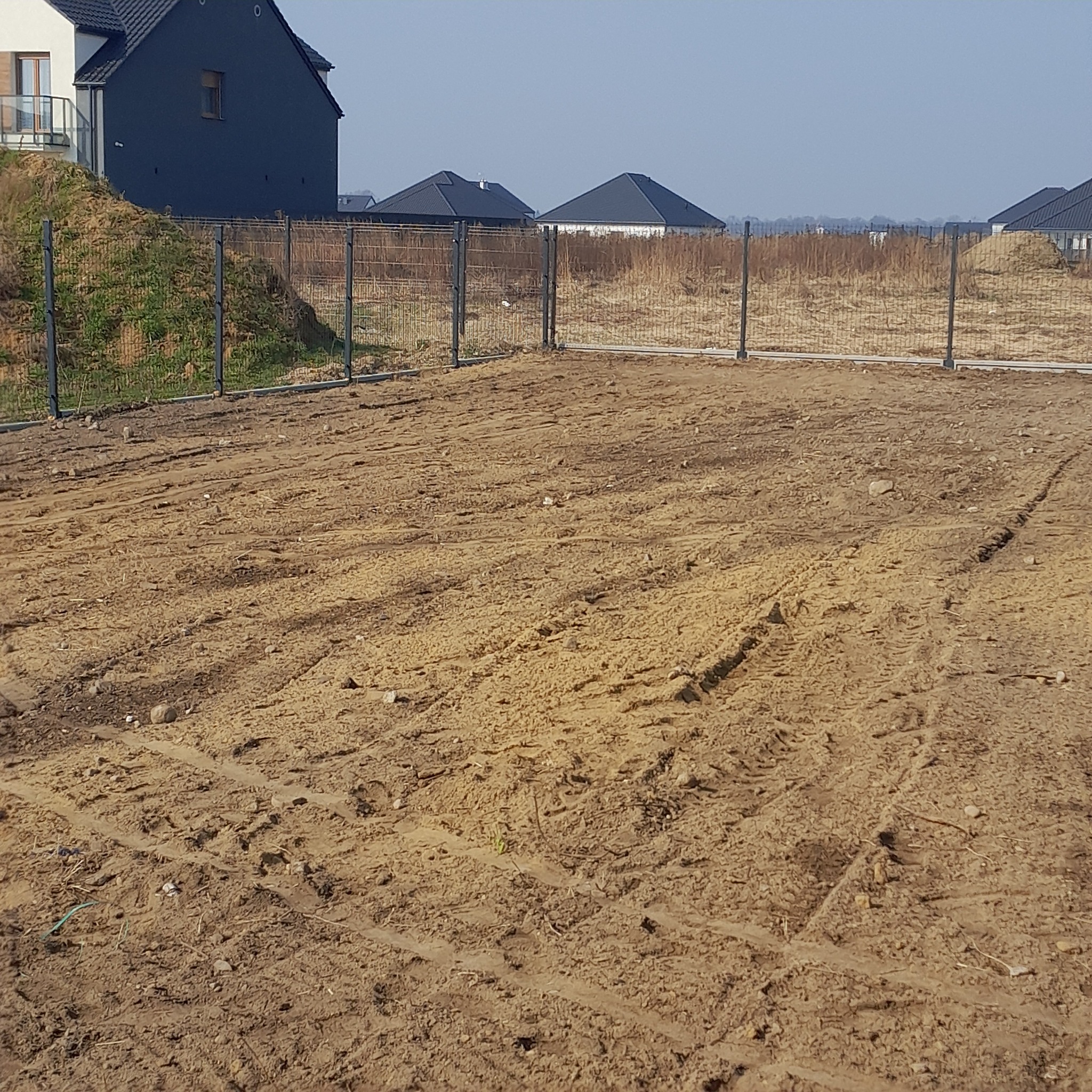 Nowoczesne ogrodzenie panelowe w kolorze grafitowym, otaczające teren budowy z widocznymi śladami opon. W tle domy jednorodzinne i błękitne niebo.